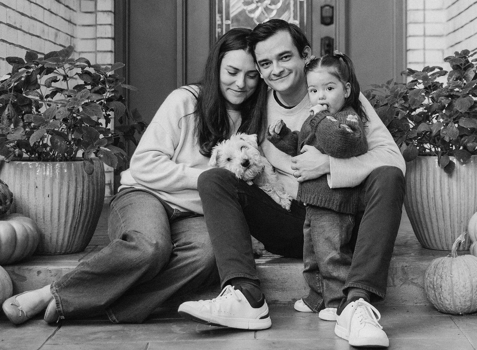 A black-and-white photo of a family sitting on steps outside their house, accompanied by pumpkins and plants. The family includes a woman, a man, a young girl, and a small dog. The woman and man are sitting close, with the woman resting her head on t