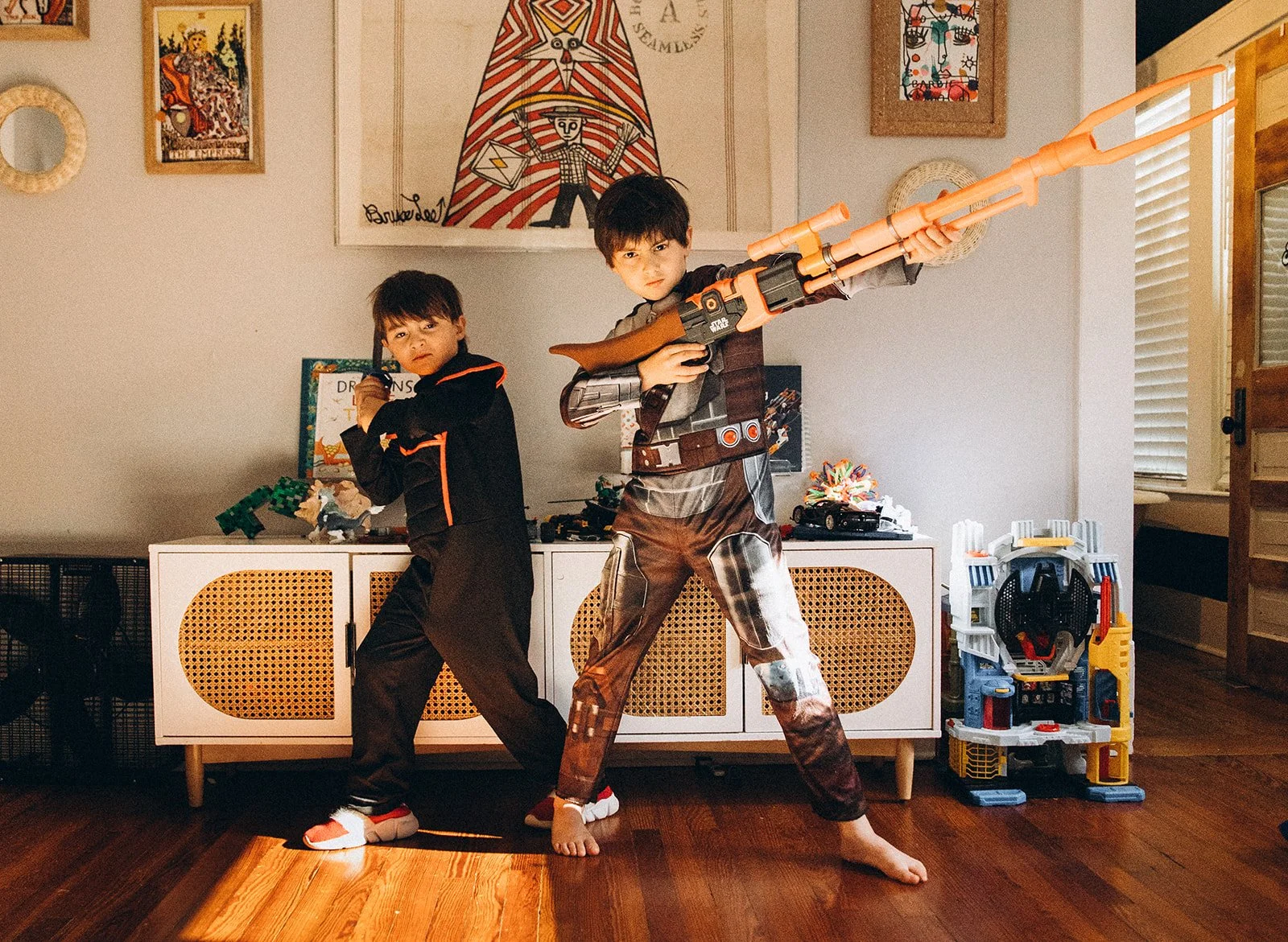 Two children inside a room, one holding a large toy rifle and the other holding a smaller toy, standing in front of a white cabinet with toys and drawings on the wall behind them.