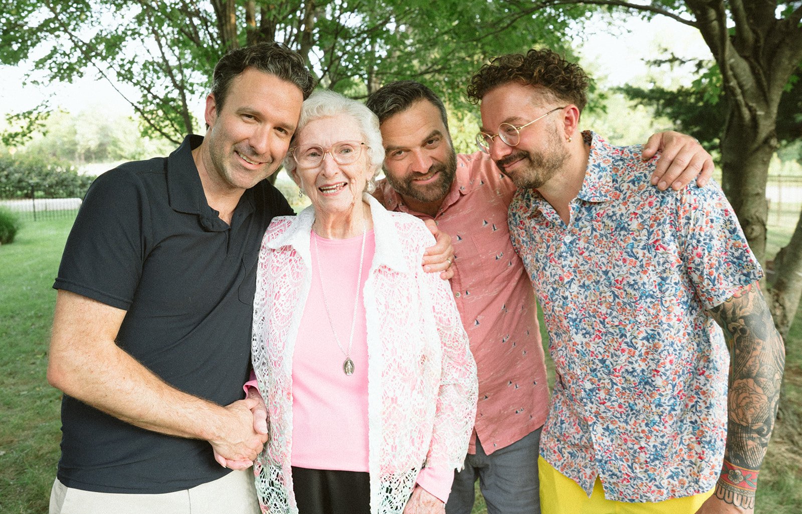 Four men and an elderly woman smiling outdoors in a park with green trees in the background.