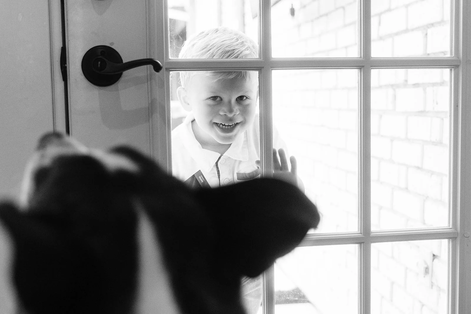 A young boy smiling and waving through a glass door while a dog is inside, looking at him.