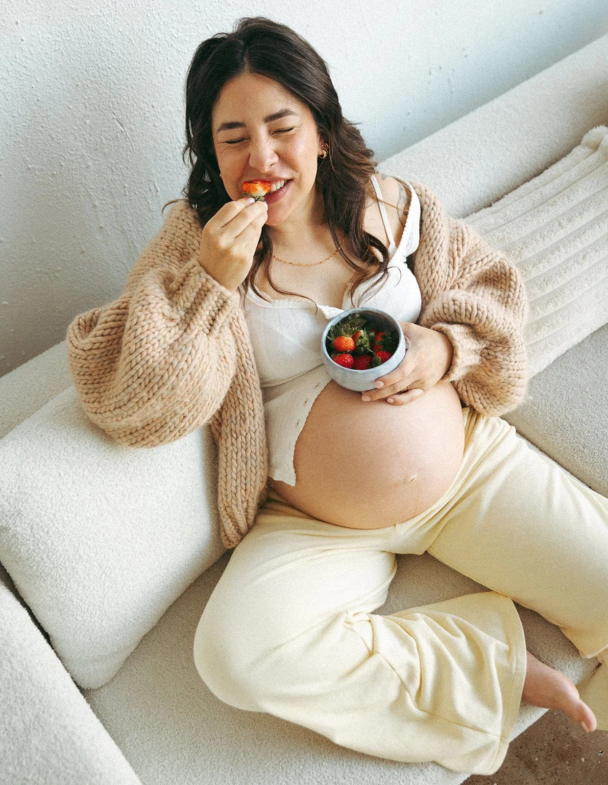 Pregnant woman sitting on a beige couch, eating strawberries from a bowl, with her head tilted back smiling.