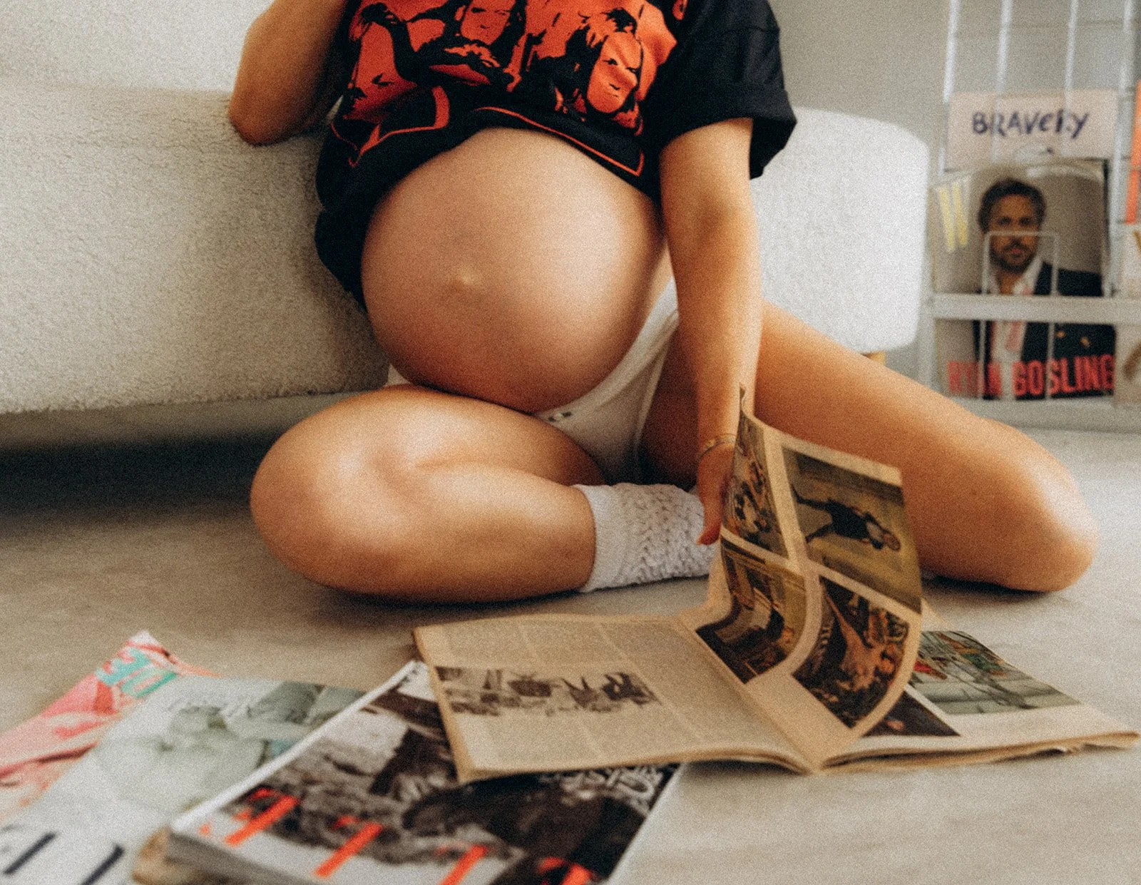 Person sitting on the floor next to a couch, reading a magazine with other magazines nearby, wearing a black and red graphic t-shirt, shorts, and white socks.