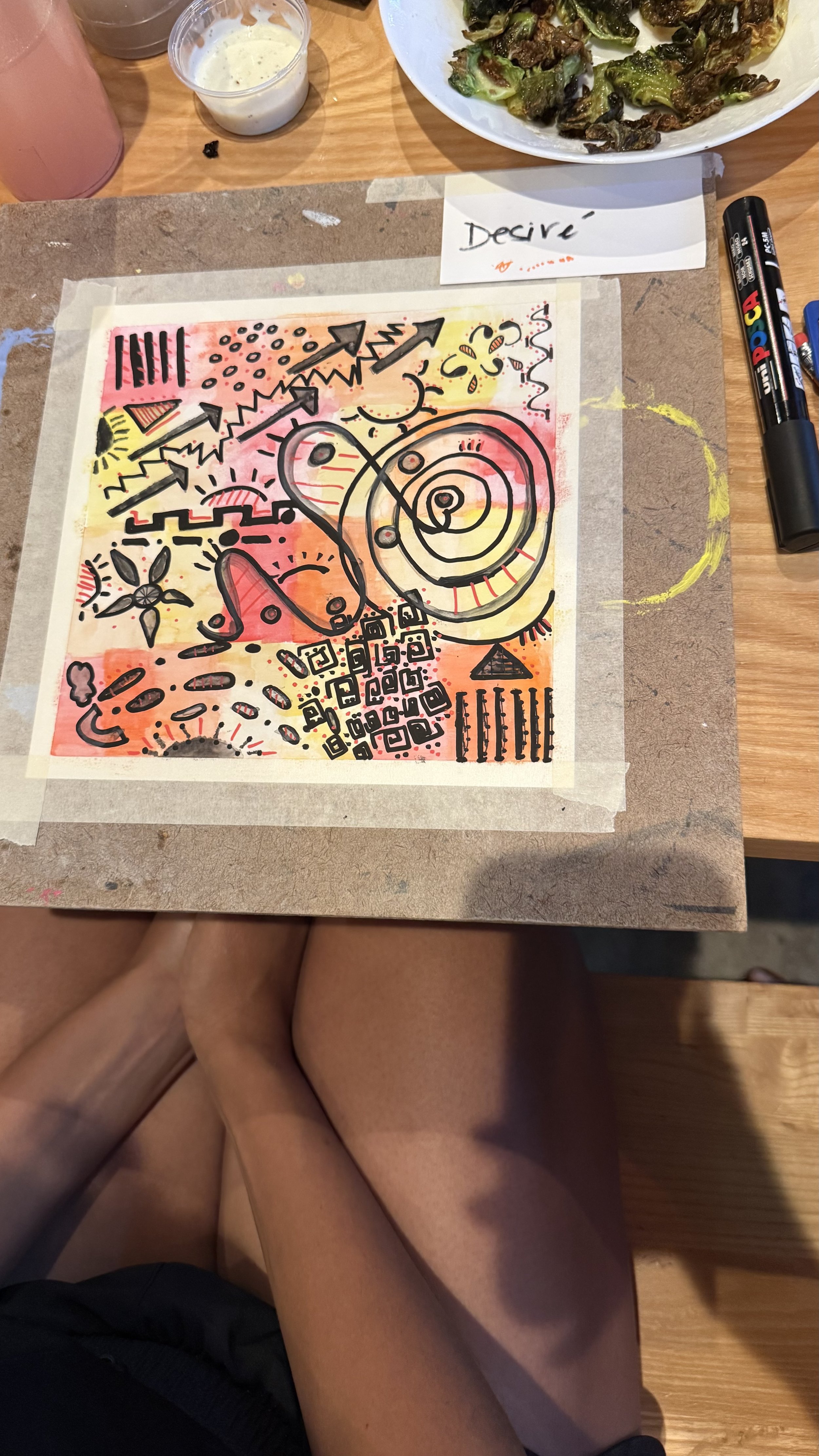 A colorful abstract art piece with various shapes, lines, and patterns in black ink on watercolor background in shades of pink, yellow, and orange. Items above include a plate of roasted Brussels sprouts, a cup of creamy sauce, and two bottles of markers.