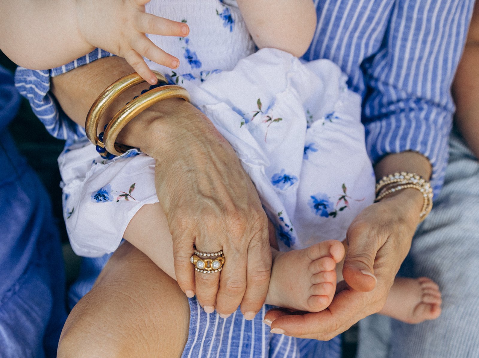 Close-up of an older woman's hands gently holding a young child's foot, with the woman wearing gold jewelry and the child in a white dress with blue floral pattern.