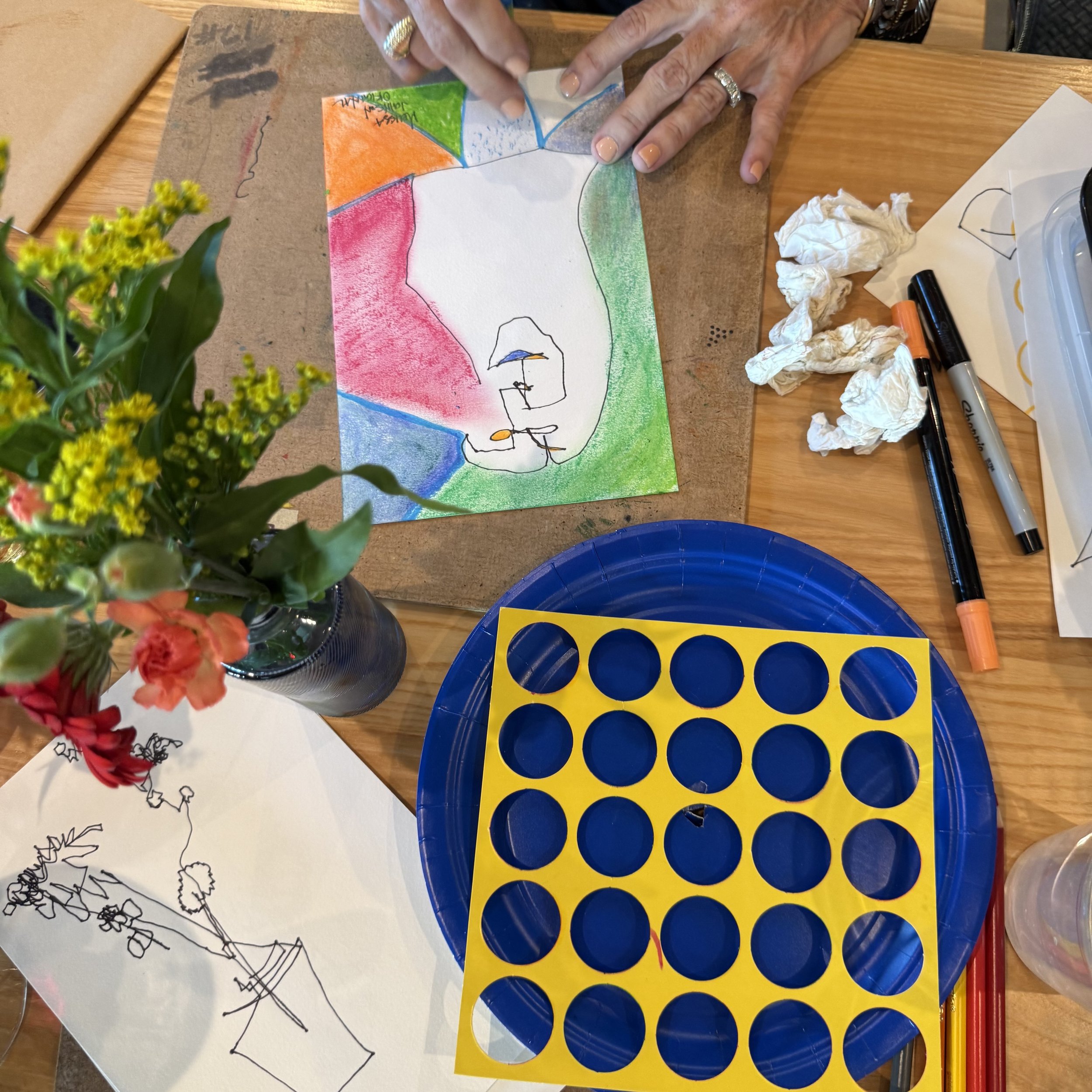 Colorful drawing with a mountain and person with an umbrella, surrounded by various art supplies including pens, tissue paper, and a blue plastic tray.