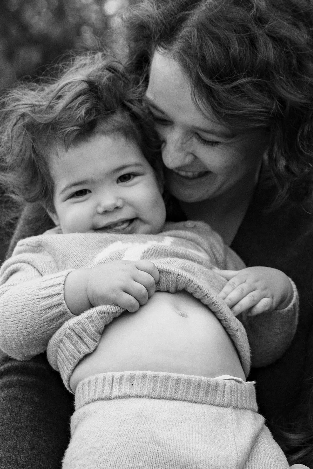 A woman and young child sharing a joyful moment, with the child revealing a swollen belly and smiling at the camera, in black and white.