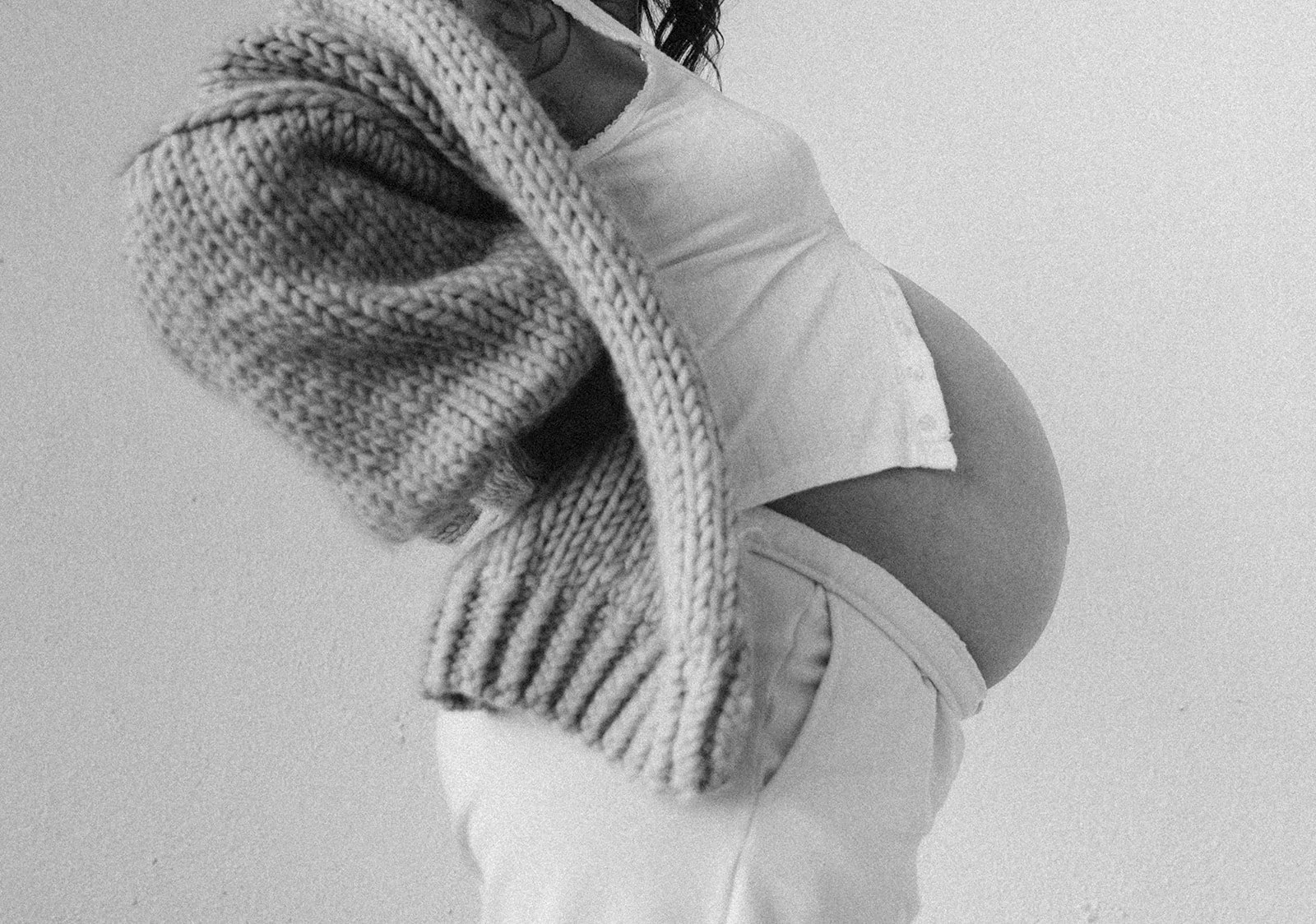 Black and white photo of a pregnant woman in a white outfit, gently holding her abdomen, with a chunky knit scarf wrapped around her shoulders.
