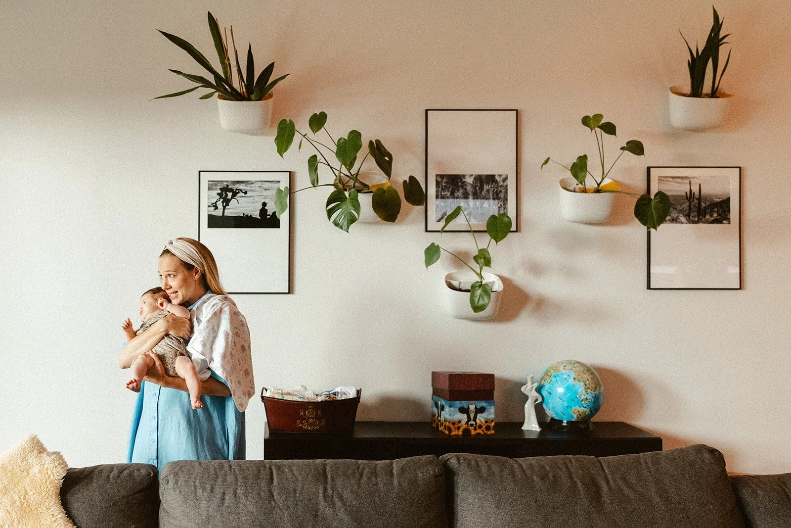 A woman holding a baby inside a living room with framed pictures, potted plants on the wall, and a shelf with decorative items, including a globe and a small statue.