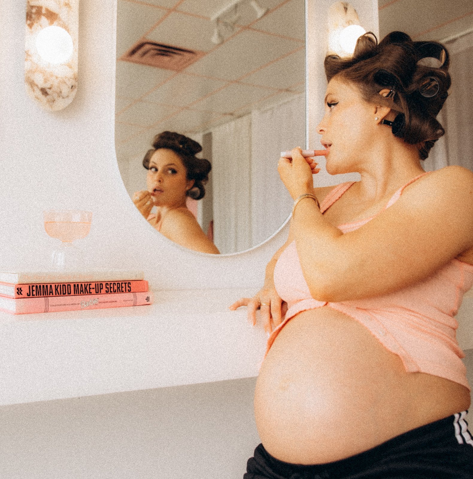 A pregnant woman with curled hair and wearing a pink camisole and black sweatpants, applying lipstick while looking in a round mirror. On the white shelf below the mirror, there are two pink books, one titled "Jemma Kidd Make-Up Secrets."