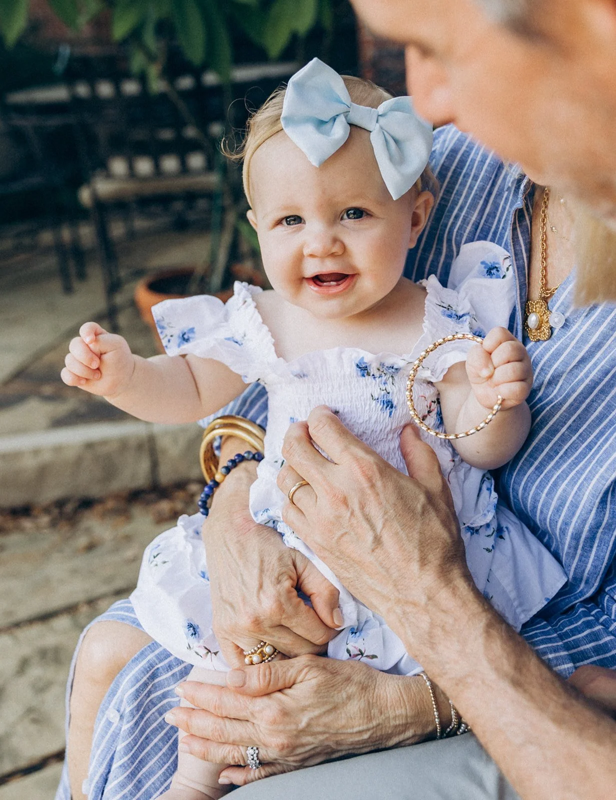 A young girl with a blue bow in her hair, wearing a white dress with blue floral patterns, is being held by a woman with a striped shirt. They are outdoors, and the girl is smiling and making a fist.
