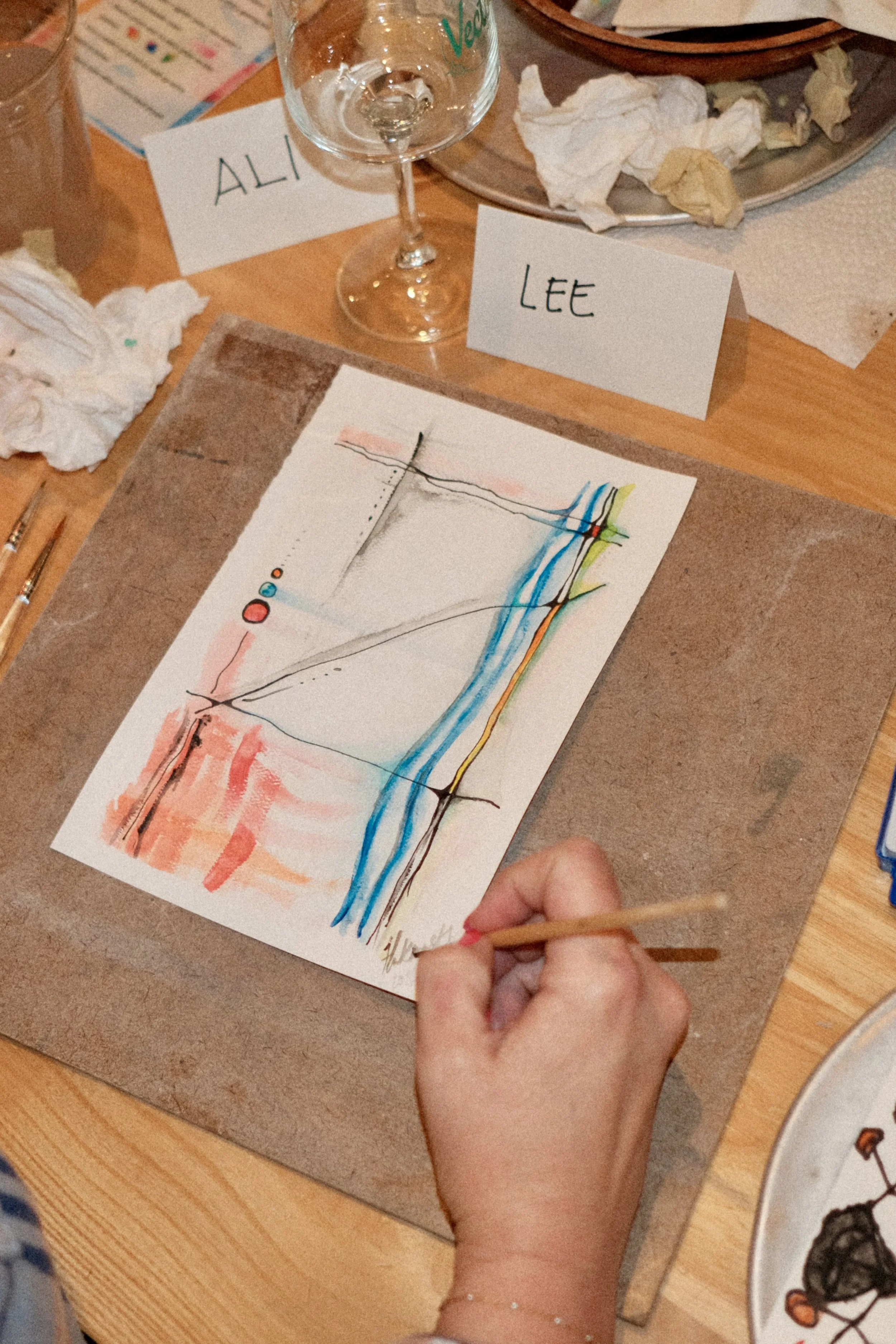 A person is creating an abstract watercolor painting with various colors including pink, orange, blue, green, black, and red on a white sheet of paper. The person's hand is holding a small paintbrush, and they are wearing a bracelet. On the table, there are crumpled tissues, a wine glass, and name cards with the names "Alicia" and "Lee".