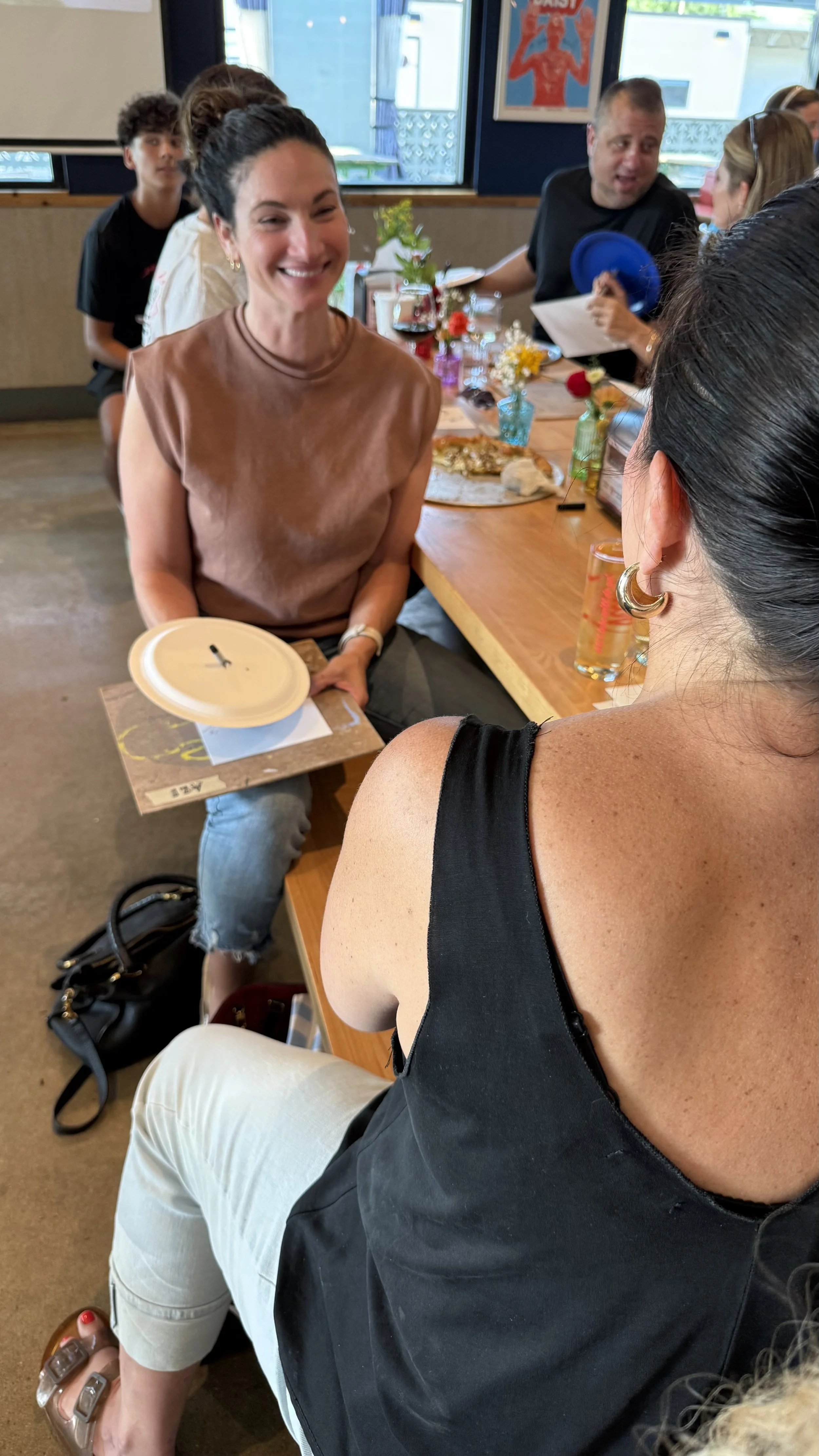 People seated at a restaurant table, engaging in conversation. The woman in the foreground is smiling, holding a plate, and wearing a brown sleeveless top. The table has colorful decor, flowers, and food. In the background, a man is looking at a woman across the table, and a young person is seated behind them. The decor includes vibrant flowers and artwork on the wall.