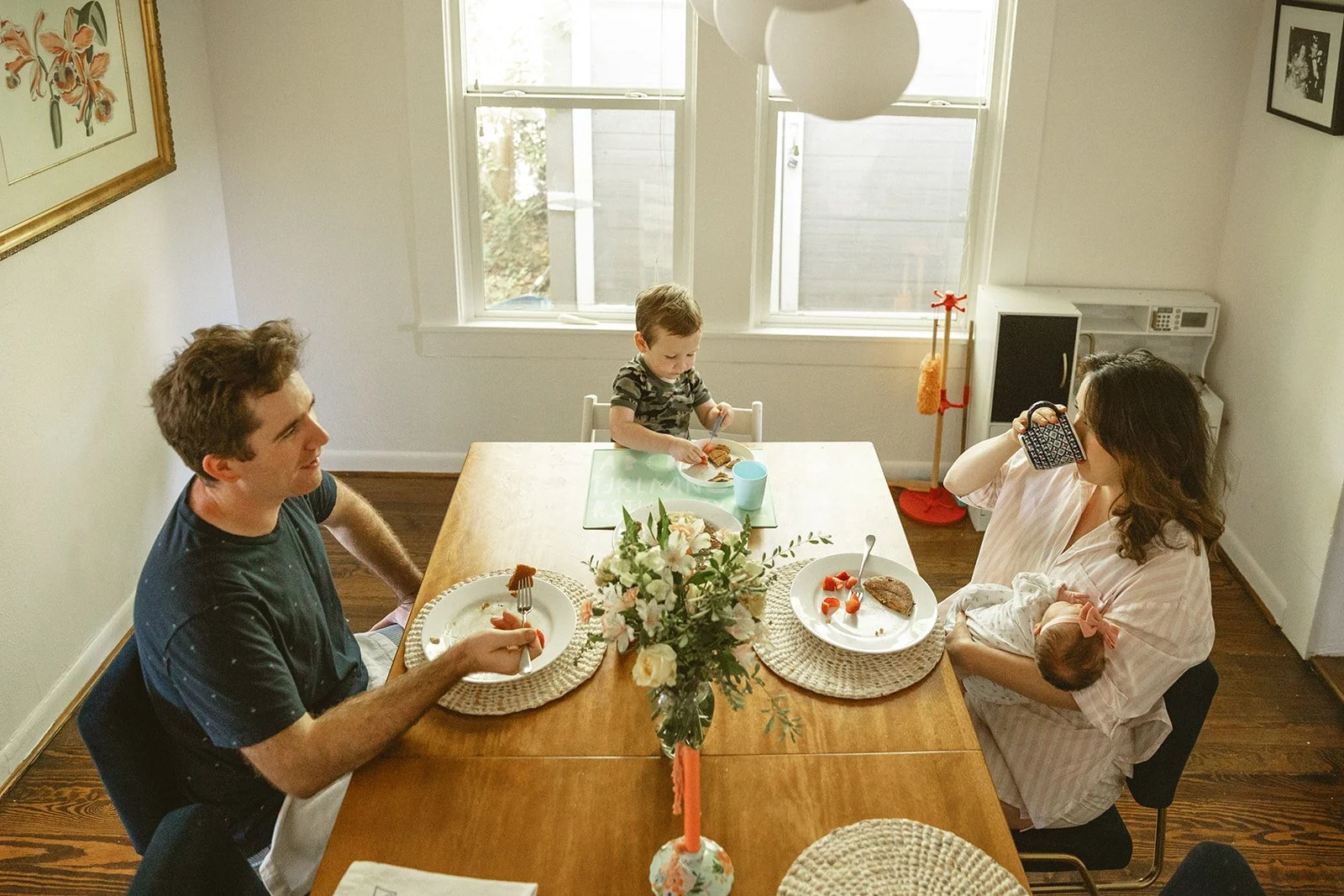 A family of four at a dining table, with a man, woman, and two children, eating breakfast in a sunlit room. The woman is holding a baby, the man is holding a fork, and the boy is eating at the table. There is a vase with flowers in the center of the 