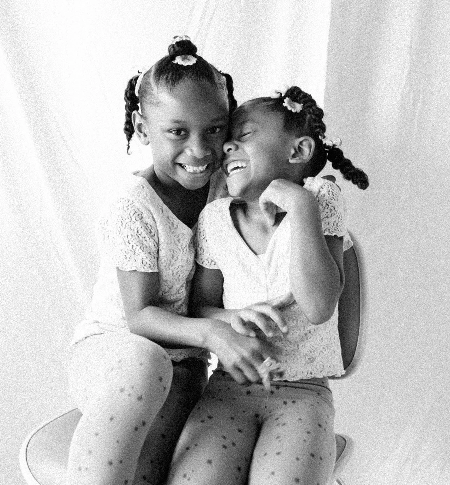 Two young girls with braided hair and polka-dotted leggings smiling and laughing together, sitting closely on a chair, with one girl hugging the other.