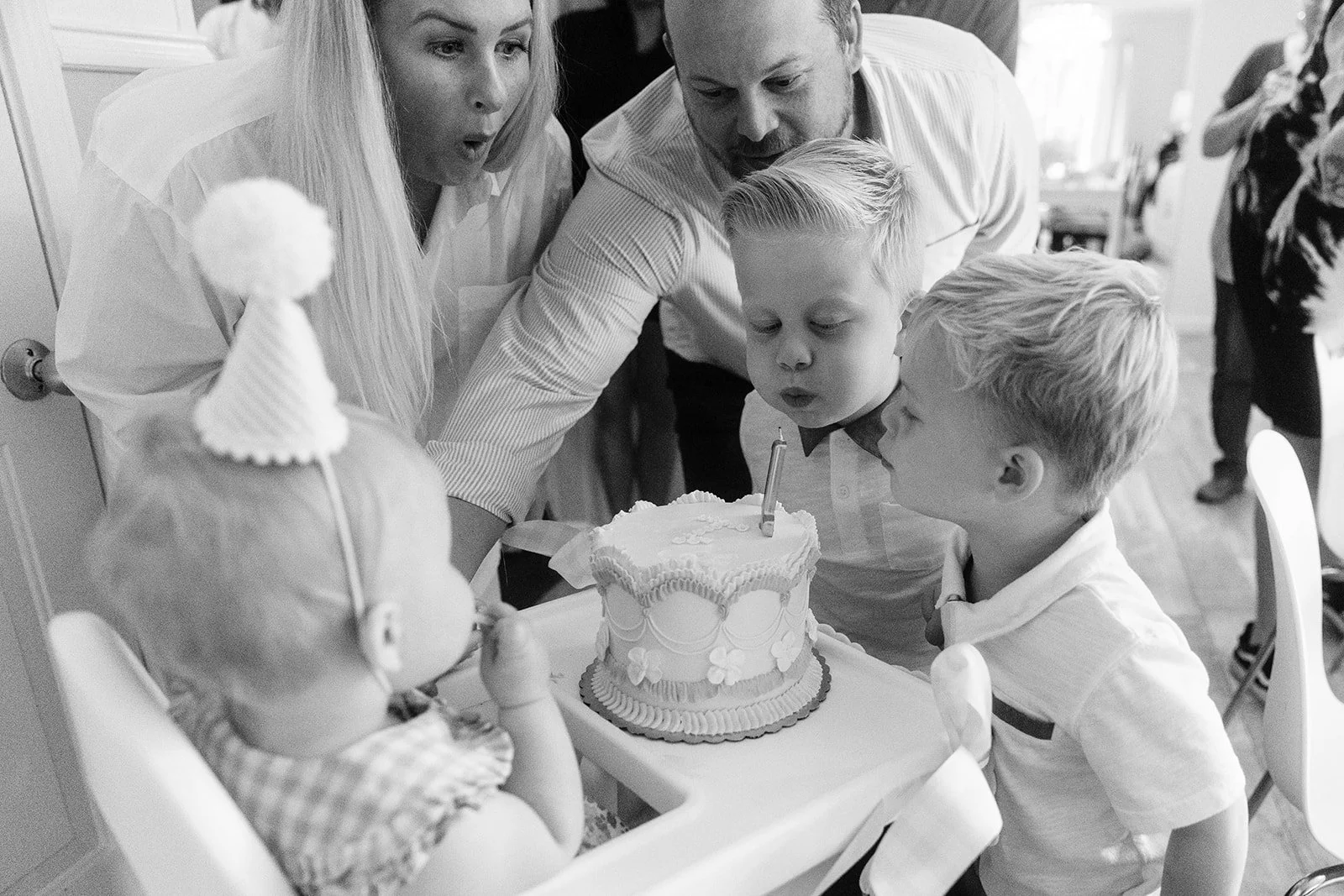 Family celebrating a child's birthday with a cake, blowing out the candles, in a home setting.