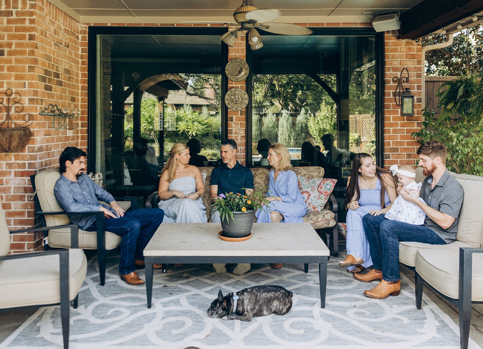 A group of seven people, including a small child, sitting on outdoor patio furniture having a conversation, with a brindle-colored dog lying down on the rug in front of them, in a backyard with greenery and a house in the background.
