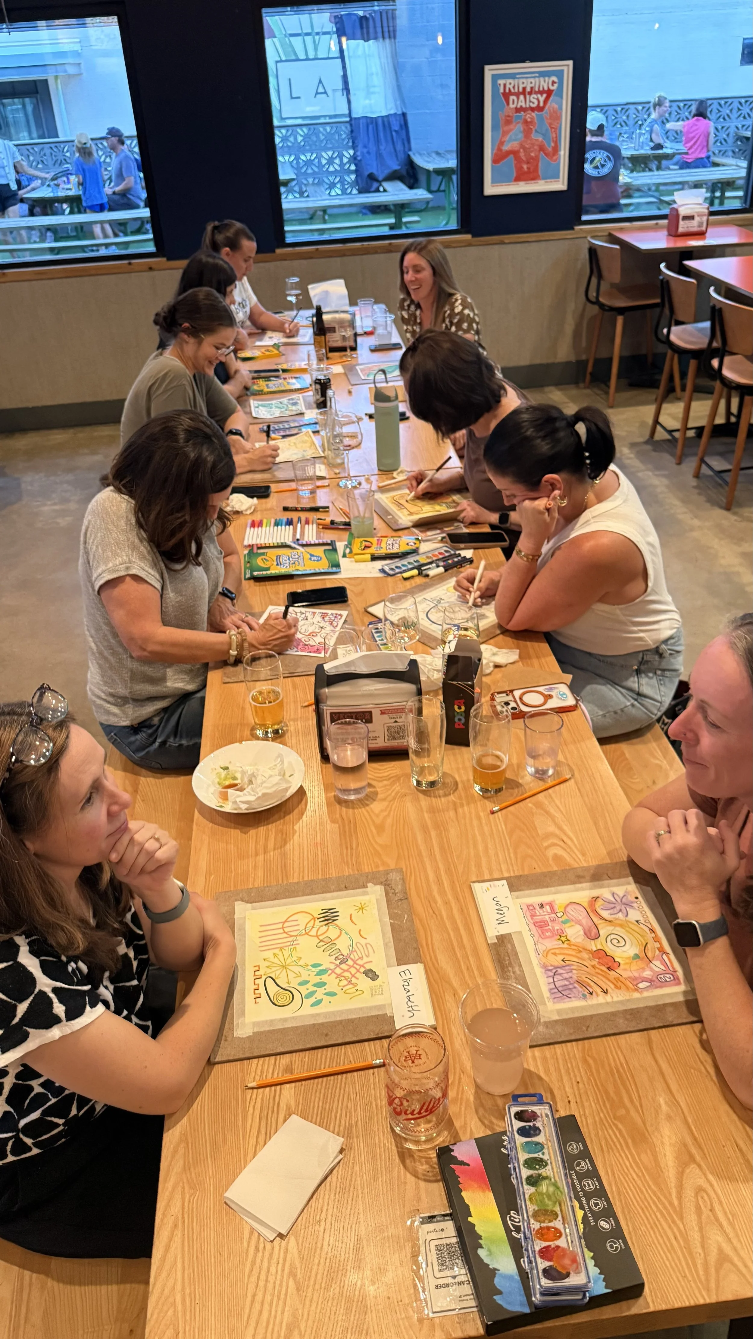 Women sitting at a long wooden table in a restaurant, coloring and drawing with various art supplies including markers, colored pencils, and watercolors.