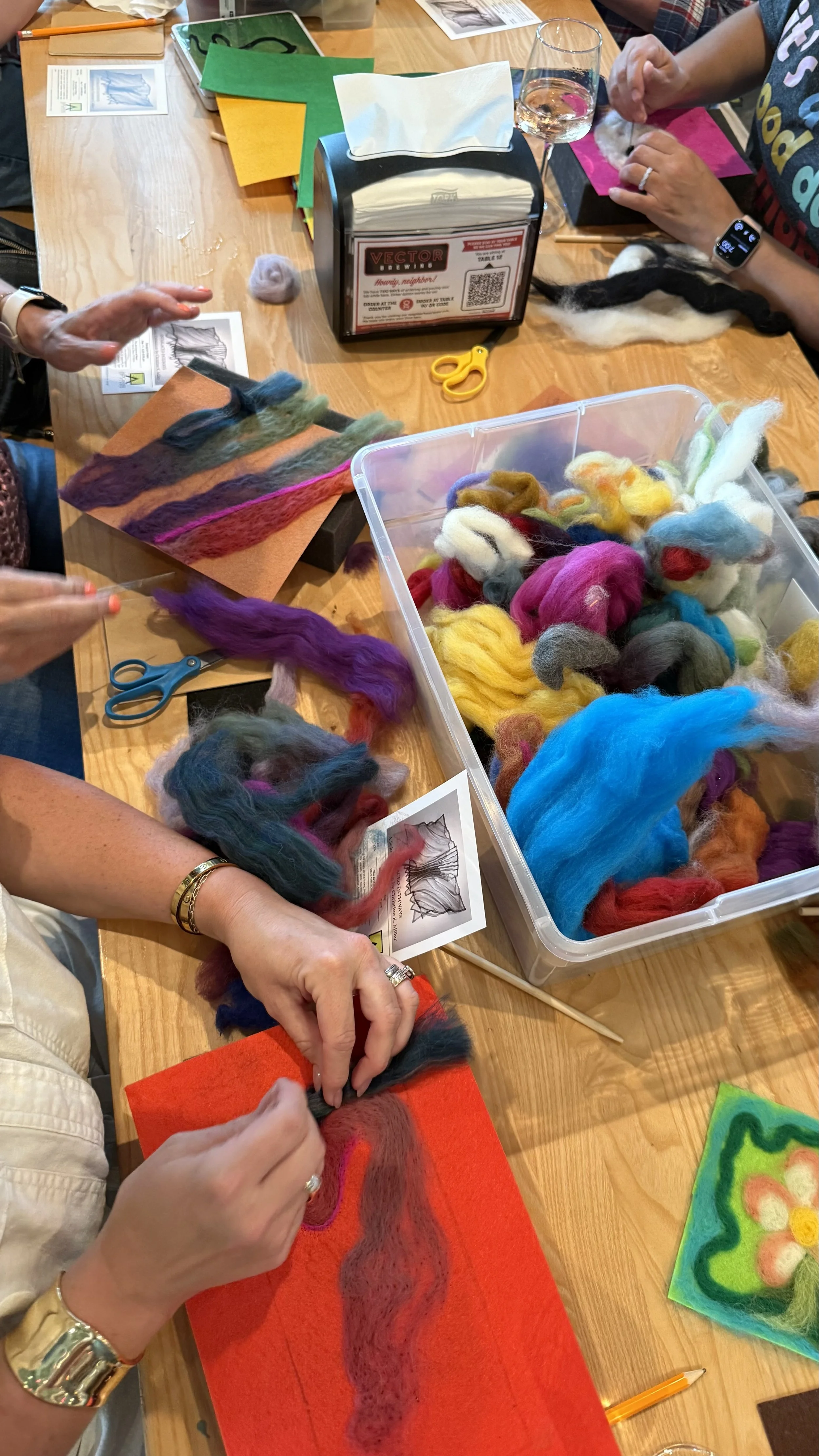 People sitting at a table working with colorful felted wool, scissors, and felting tools. They are creating felted art on felt boards, with a container of multicolored felt wool and felting instructions visible.