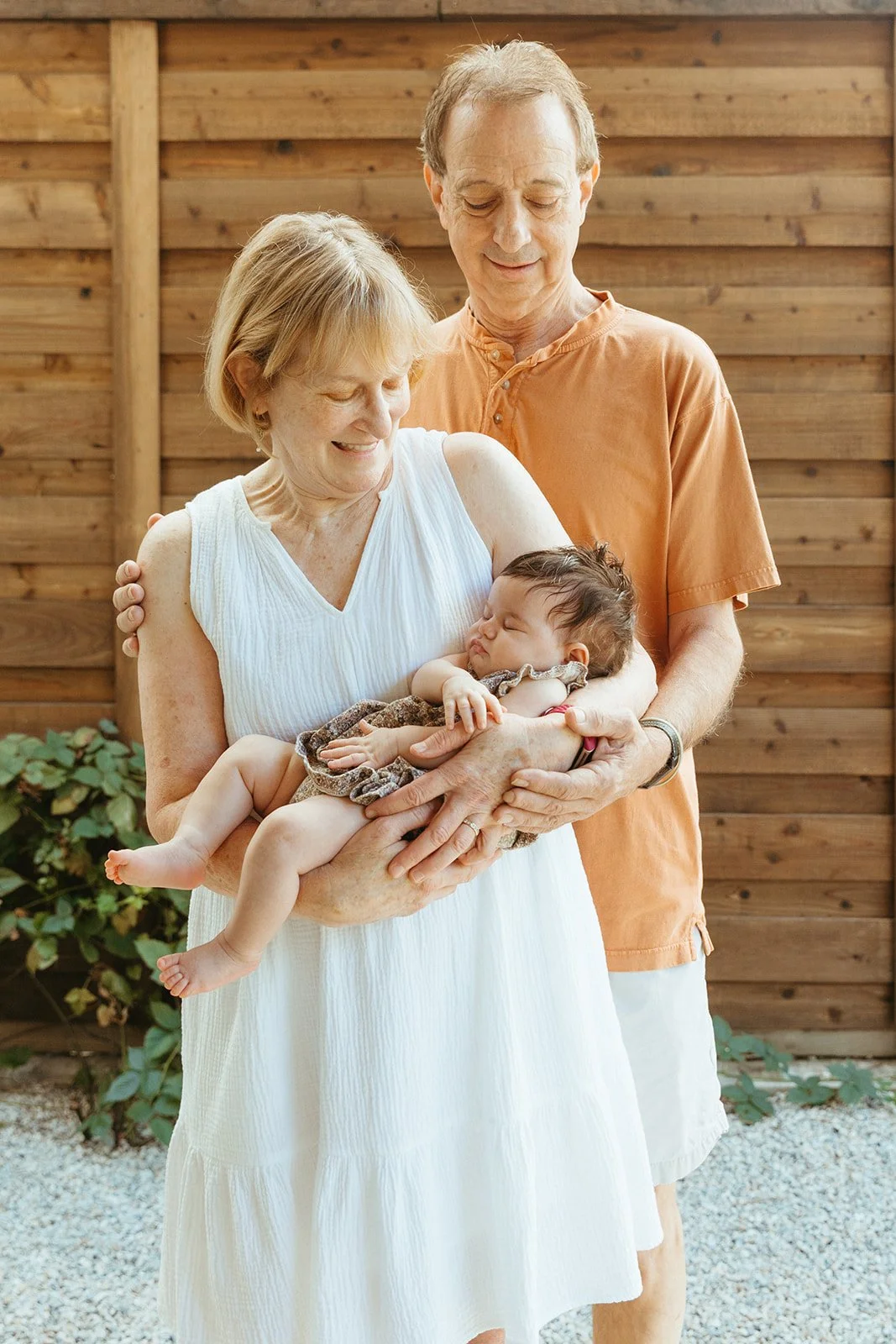 An elderly woman holding a sleeping baby with a man standing behind her, outdoors with a wooden fence in the background.