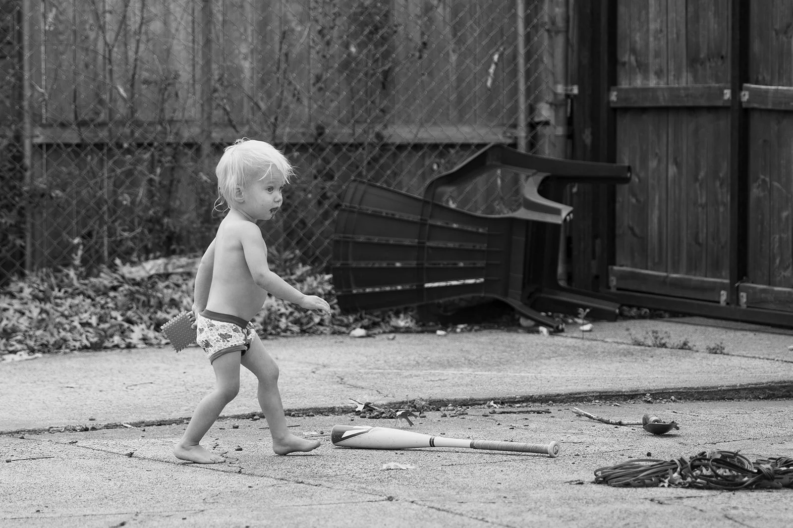 A young boy with blonde hair, wearing only underwear, is standing on a paved surface in a backyard, holding a baseball bat. There is a fallen overturned stroller in the background, along with a broom on the ground nearby. The yard is enclosed with a 