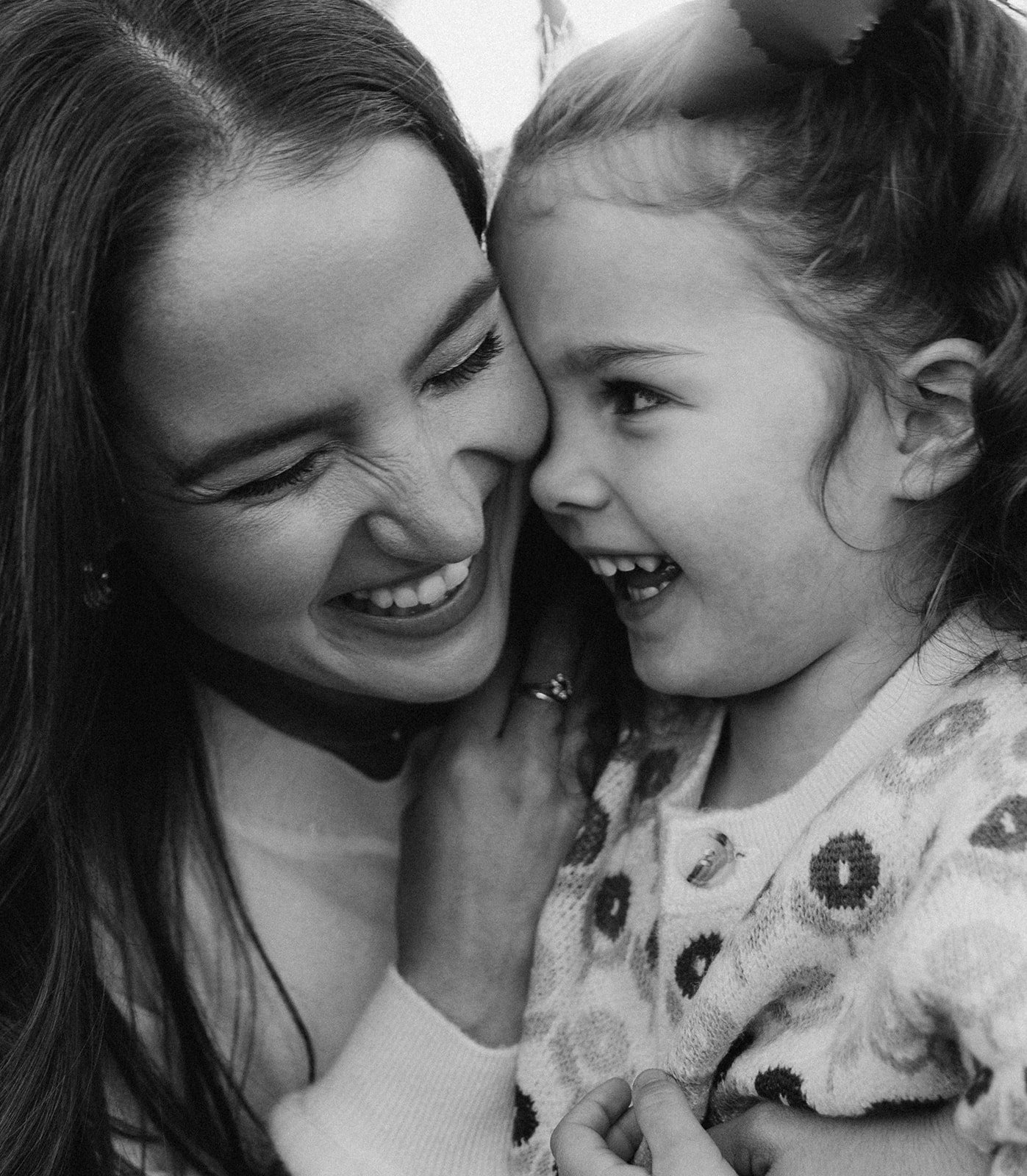 A black and white close-up photo of a woman and a young girl sharing a joyful moment, with their faces close and smiles, the woman with long dark hair and the girl with curly hair wearing a patterned sweater.
