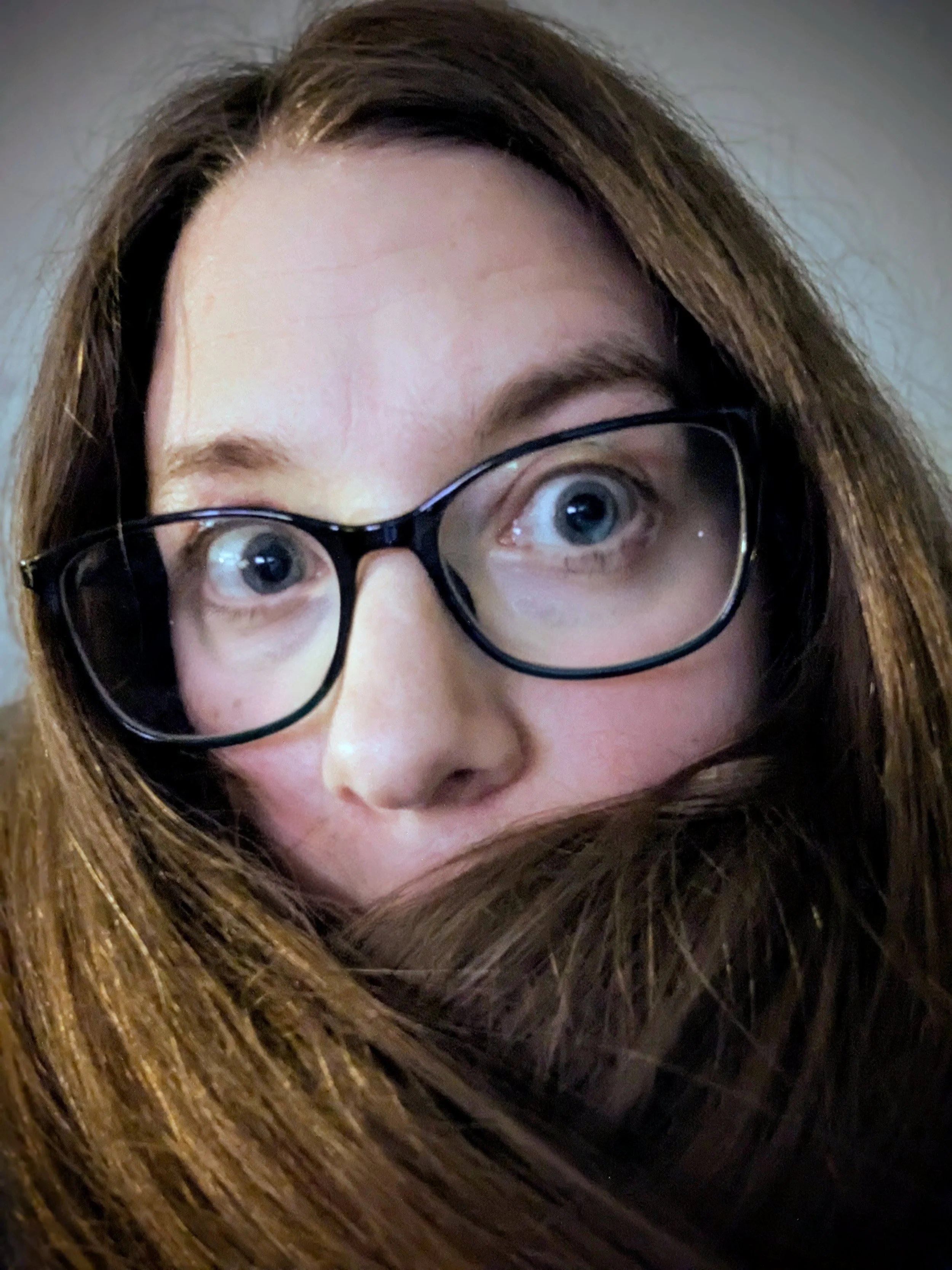 Close-up of a woman's face with brown hair and black glasses, looking up with wide eyes.