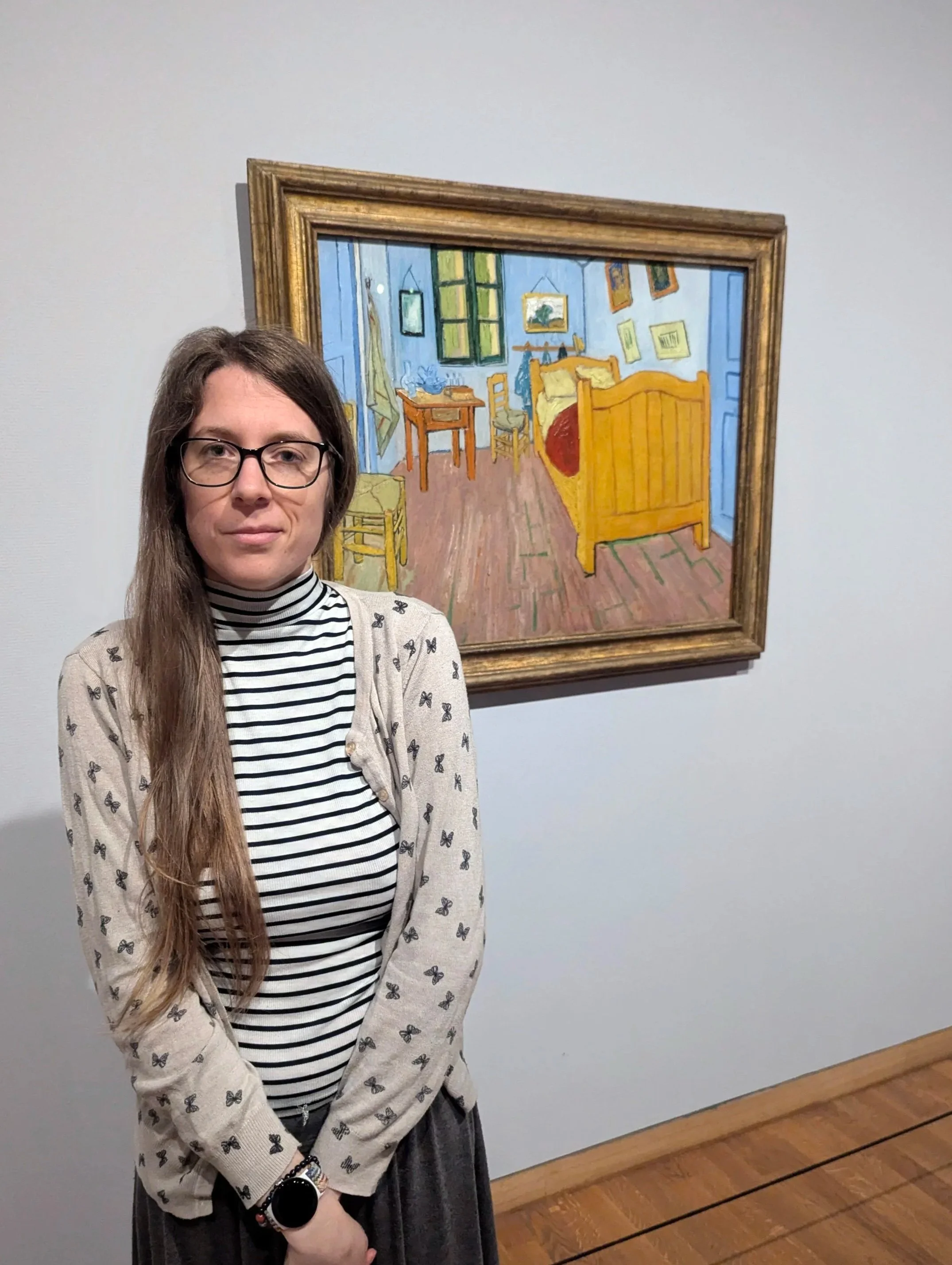 A woman with long brown hair and glasses standing in front of a framed painting of a bedroom scene, with a bed, table, chairs, and window, displayed on a white wall in an art gallery.