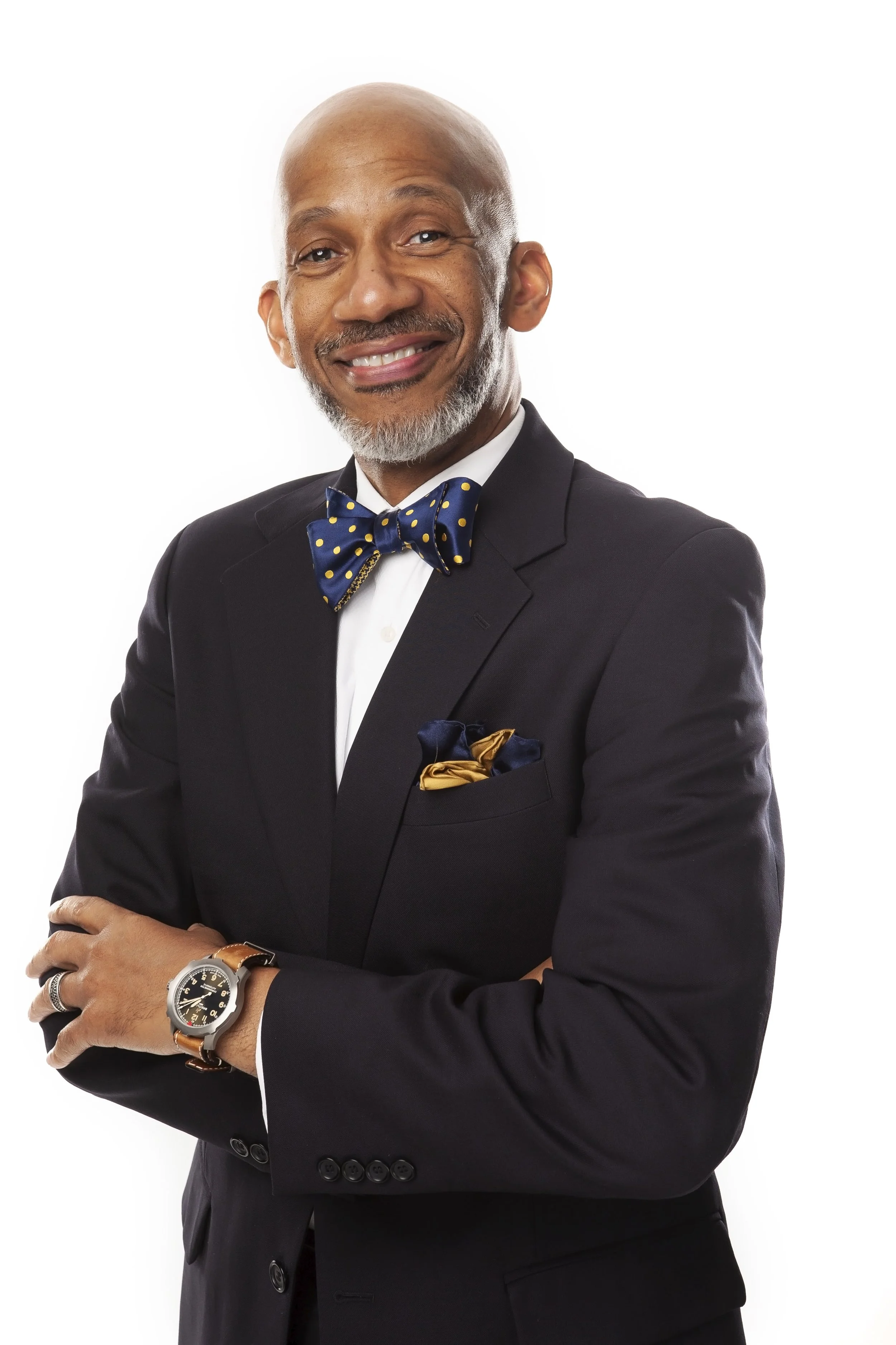 A distinguished middle-aged man with a gray beard and bald head wearing a black suit, navy blue bow tie with yellow dots, white shirt, and a gold pocket square. He is smiling and has his arms crossed, wearing a wristwatch.