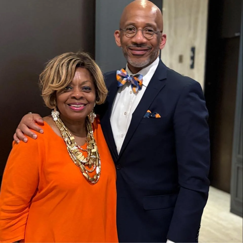 A smiling woman in an orange dress with layered gold and pearl necklaces, and a woman in a navy suit with a bow tie, standing close together in an indoor setting.