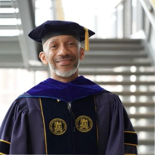 A man in a graduation gown and cap standing in front of a staircase, smiling.