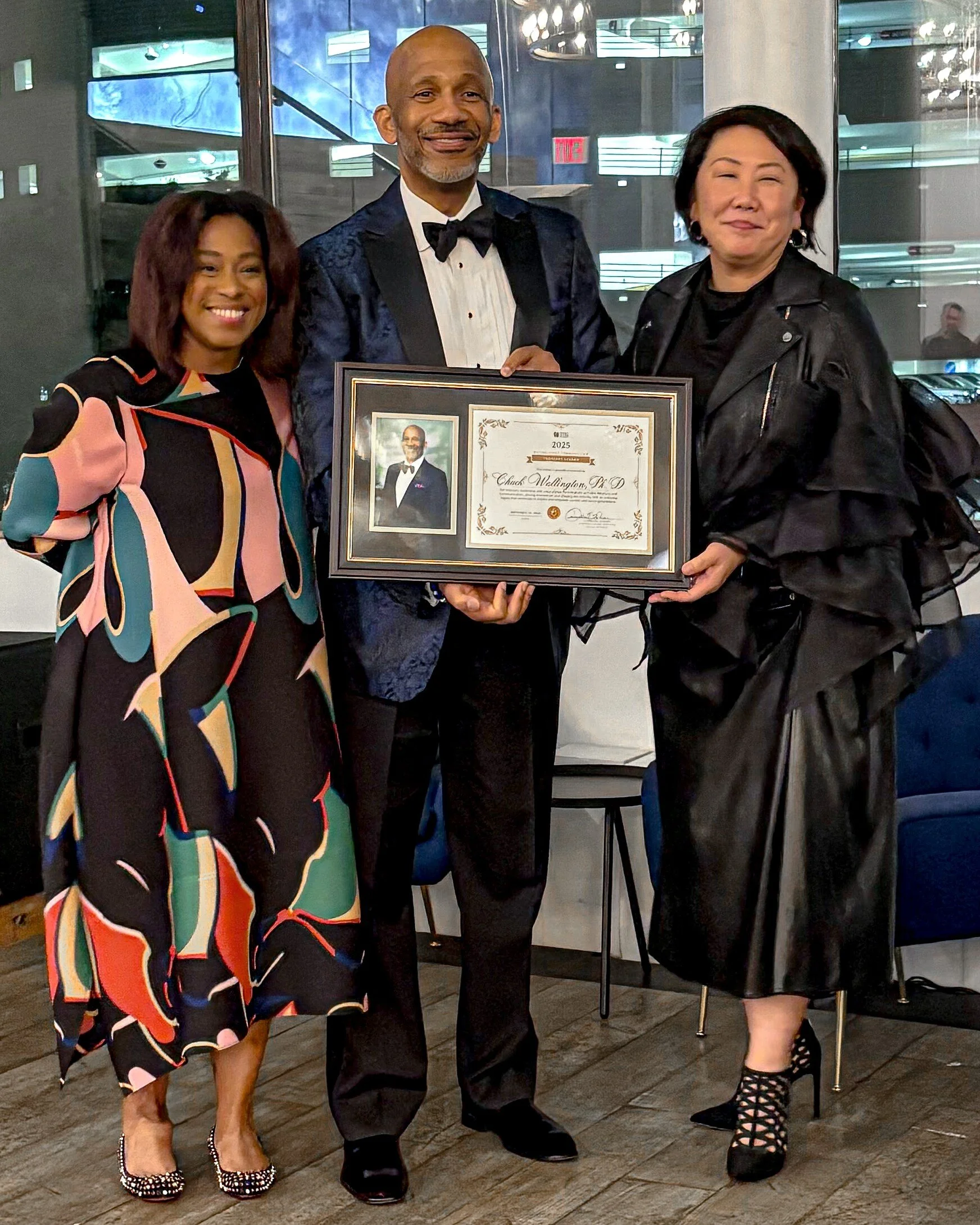 Three people standing in a room, holding a framed certificate with a photo of a man in a tuxedo. The man in the center is wearing a tuxedo and smiling. The woman on the left is wearing a colorful dress with black, pink, teal, and orange patches. The woman on the right is wearing a black leather jacket and black skirt, with lace-up heels. Behind them are large windows and chairs, suggesting an event or celebration.