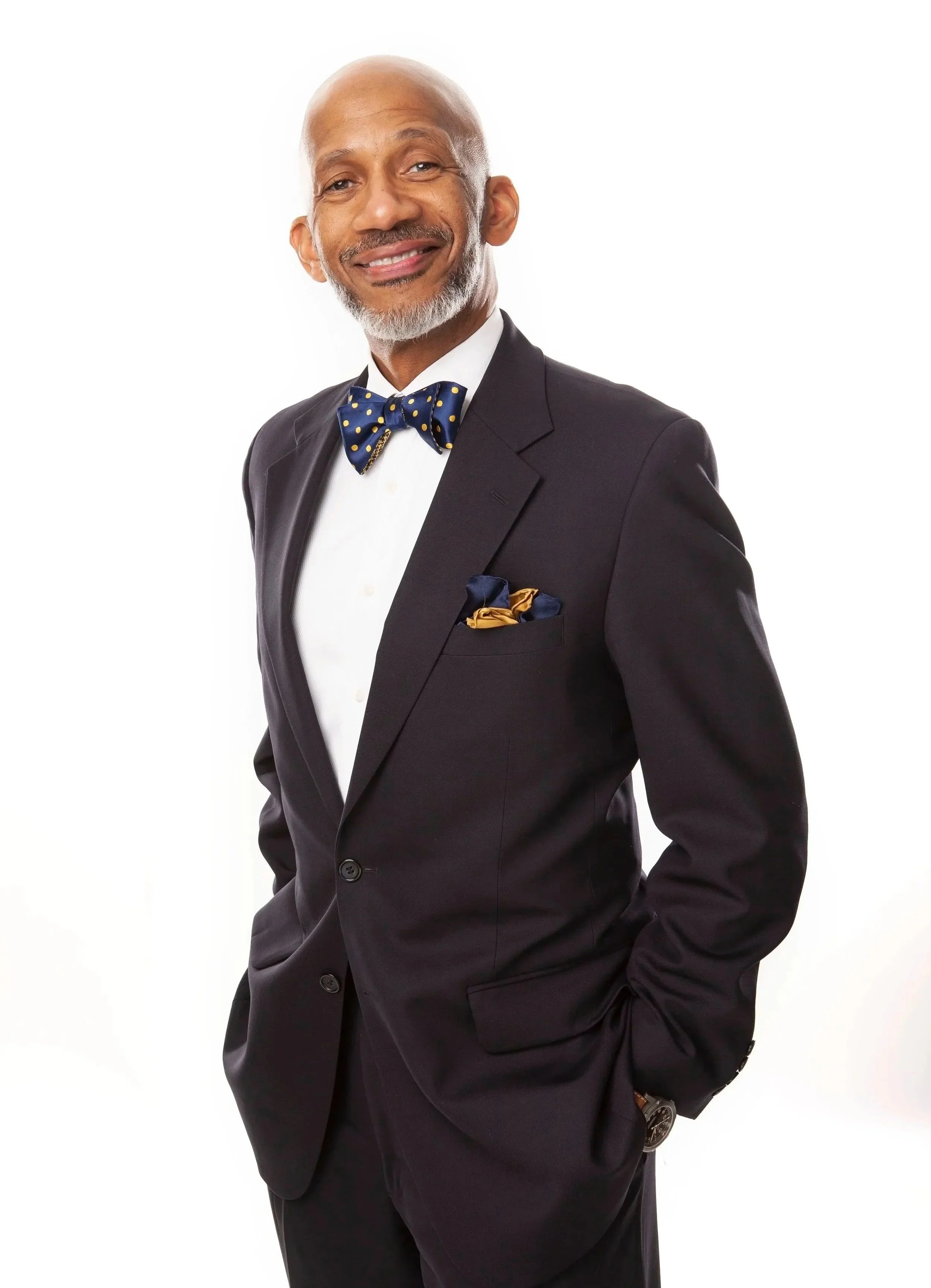 A middle-aged man with a gray beard and bald head wearing a black suit, white dress shirt, and a navy blue bow tie with yellow polka dots. He has a matching pocket square and is smiling against a plain white background.