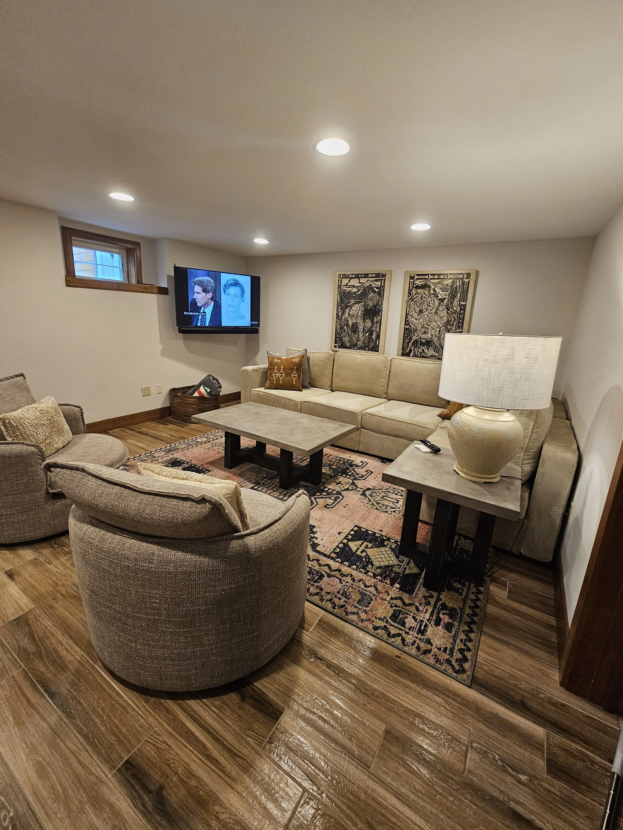 A cozy living room with beige and brown furniture, a patterned area rug, a wall-mounted TV, and framed artwork.