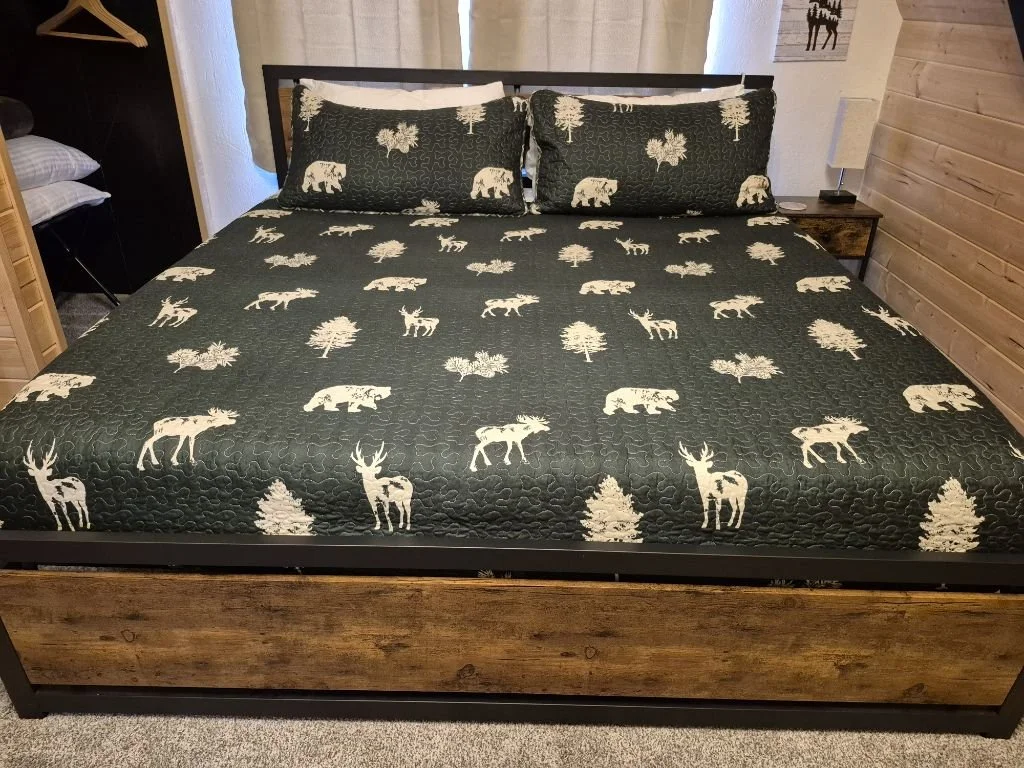 A bed with a black quilt featuring white moose and tree patterns, and two matching pillows. The bedroom has wooden walls, a nightstand with a lamp, and visible shelving.