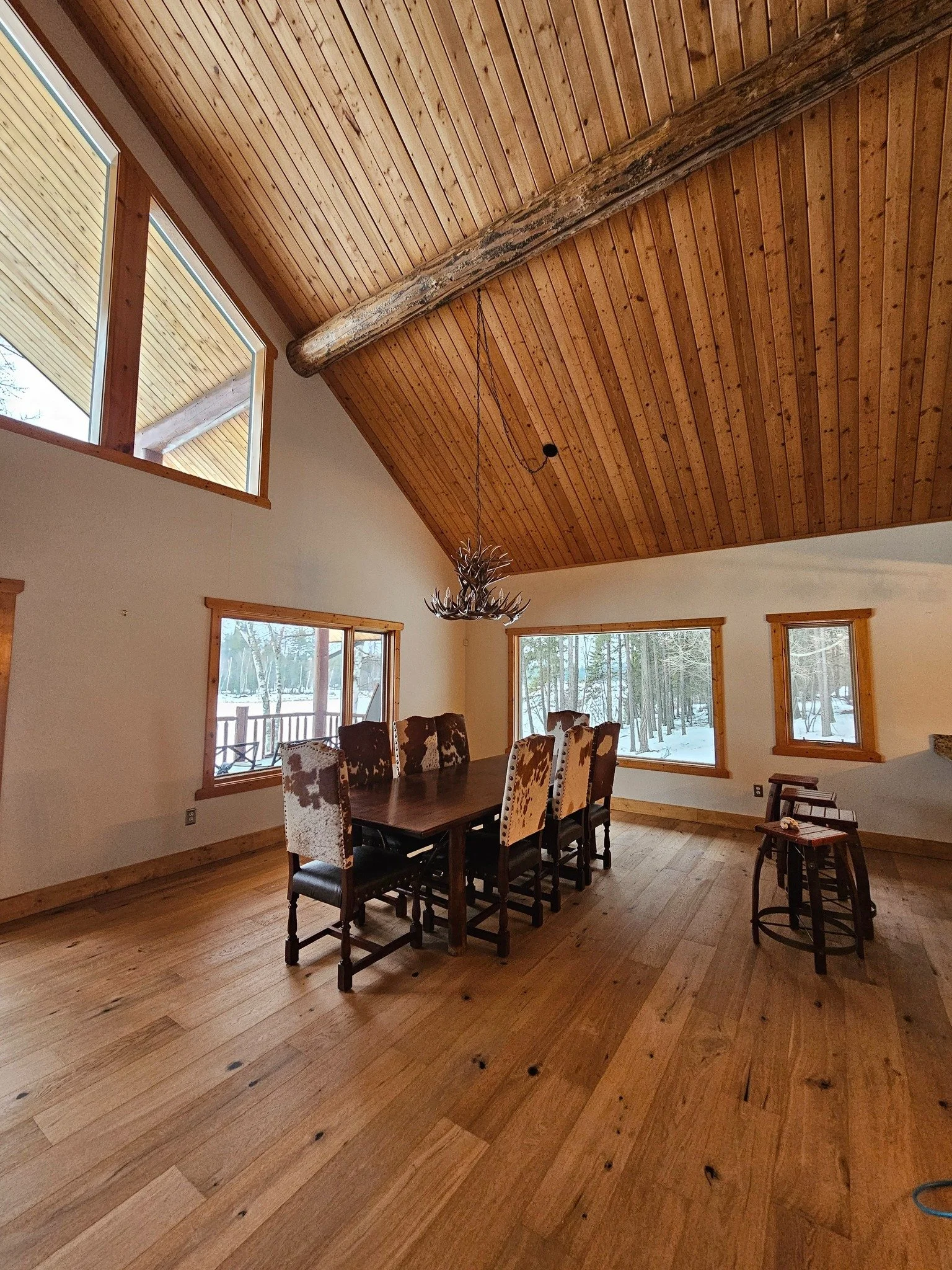 Interior view of a dining room with wooden ceiling, walls, and floor, featuring a large farmhouse-style table with cowhide chairs, a chandelier made of antlers, and multiple windows showing snow-covered trees outside.