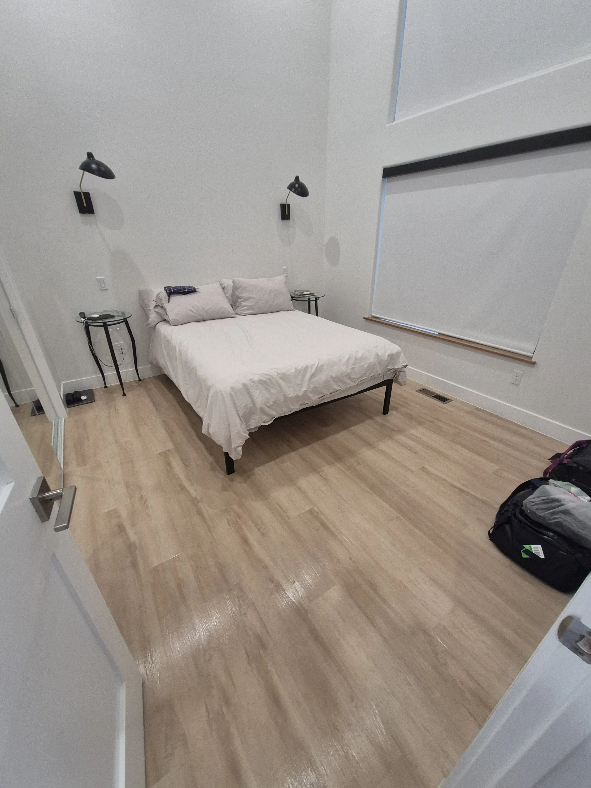A minimalist bedroom with a white bed, two black wall-mounted lamps above, two small black bedside tables, and a large window with blinds. The room has light wood flooring and a door open to the left.