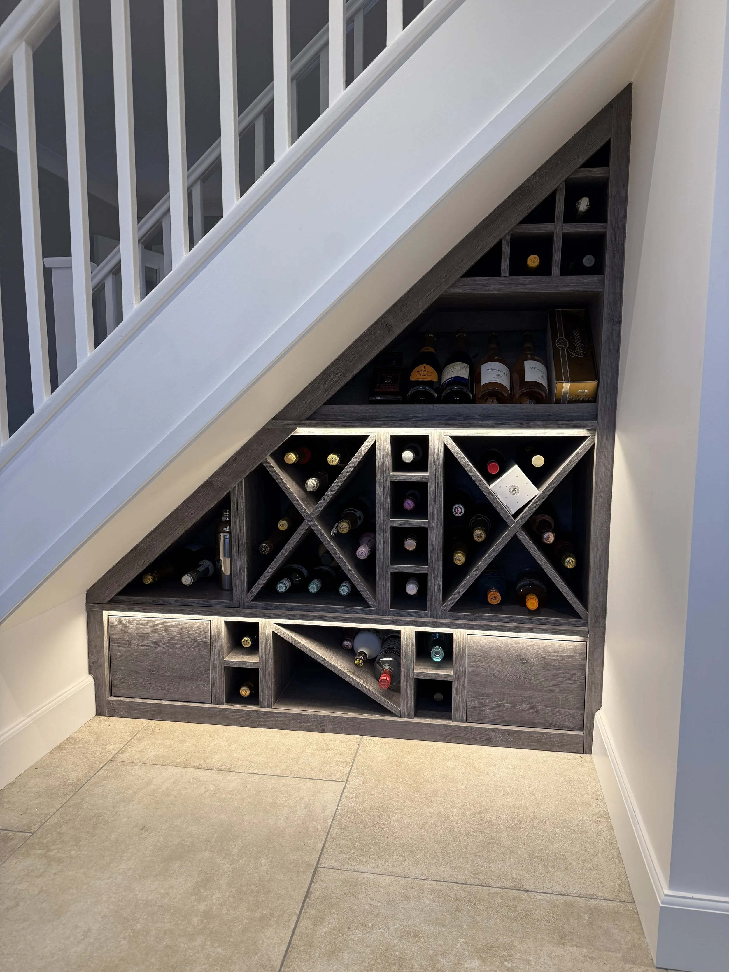 A built-in wine rack with multiple shelves and compartments, filled with wine bottles, located under a staircase with white railing, in a room with beige tiled flooring and white walls.