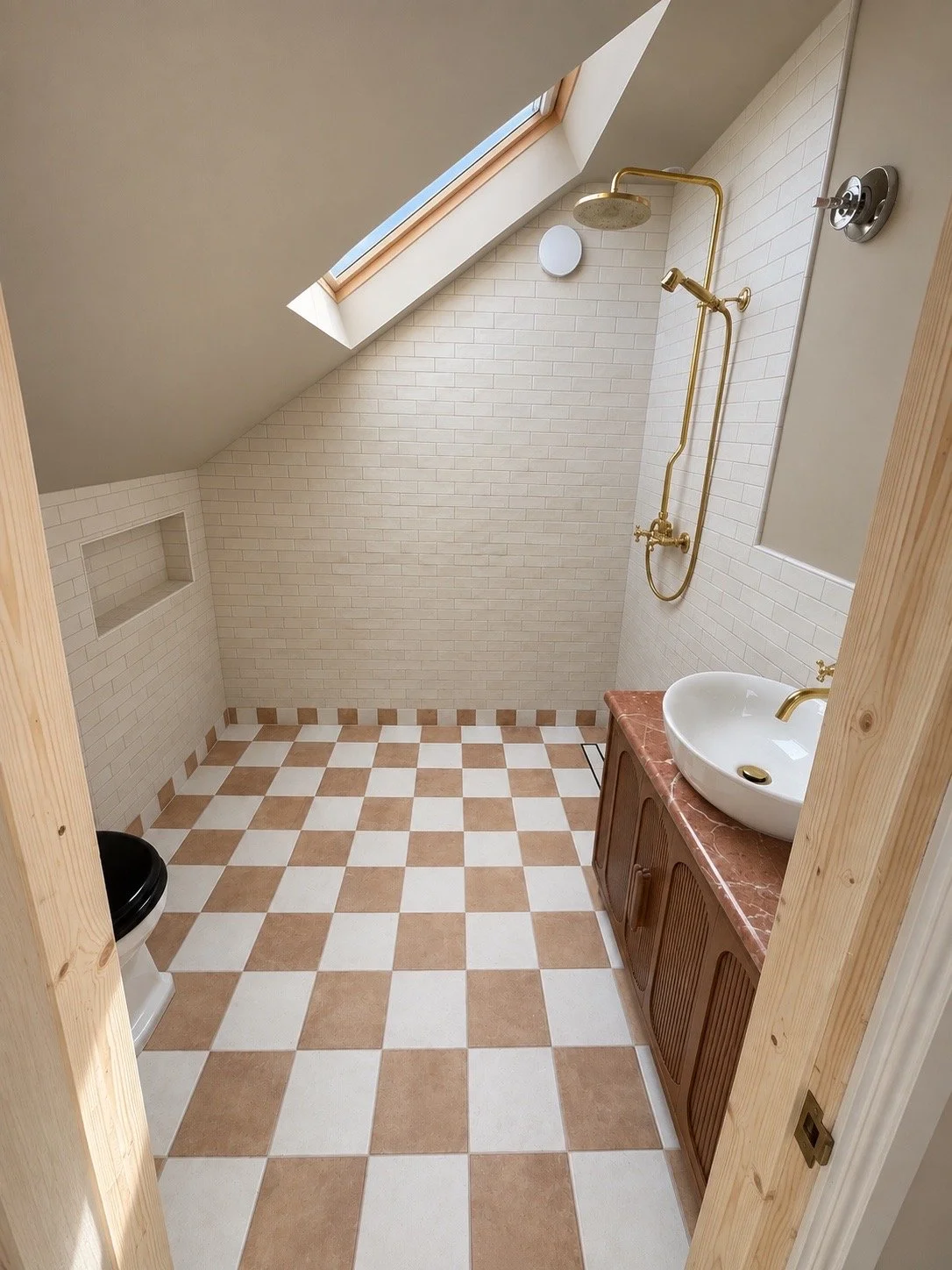 Small bathroom with sloped ceiling, skylight window, white tiled walls, checkered beige and white floor, wooden vanity with a vessel sink, shower with brass fixtures, and a black toilet.