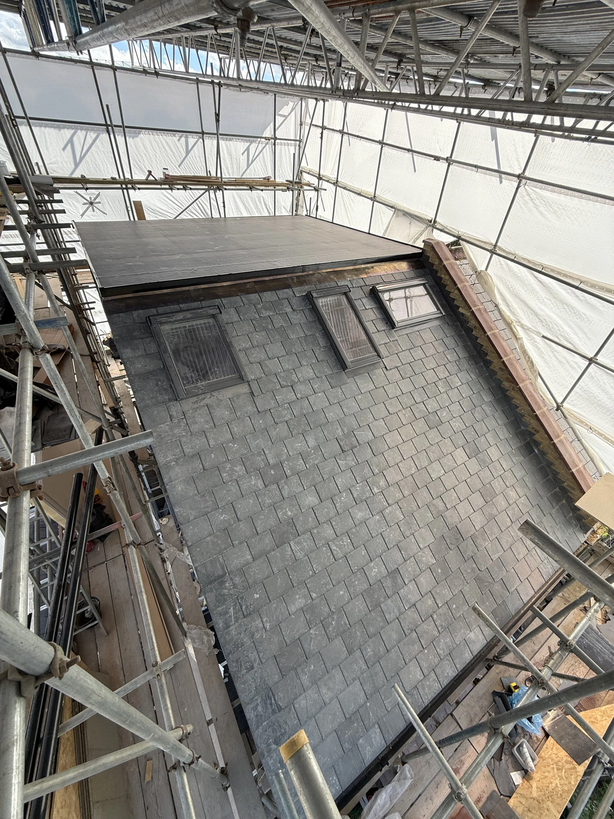 Scaffolding surrounds a house with gray brick siding and a black roof, with windows on the side of the house, and part of the roof appears to be under construction or repair.