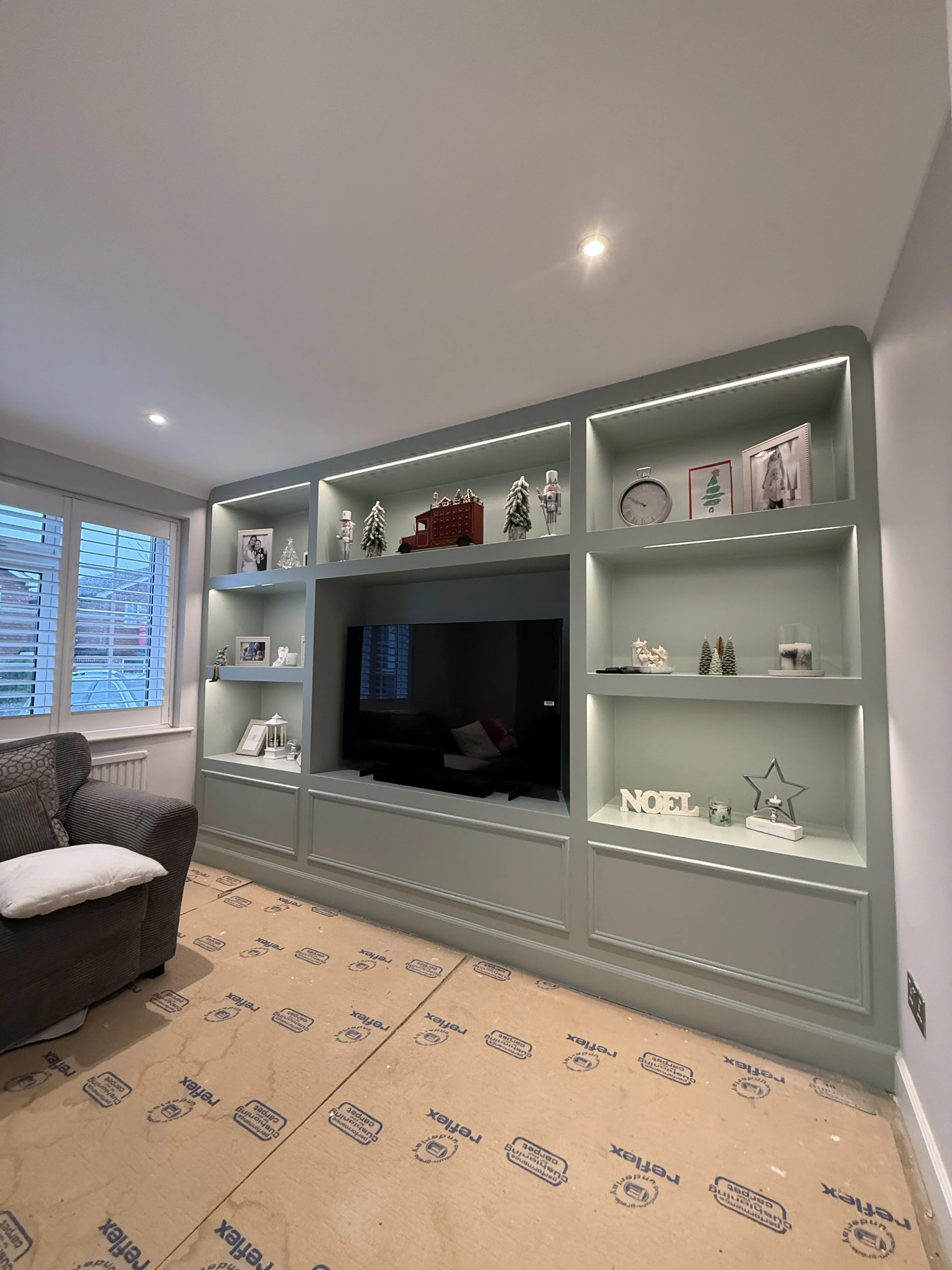 Living room with built-in shelves decorated with Christmas ornaments and photos, a large flat-screen TV, a window with blinds, a gray armchair with cushions, and cardboard flooring protection with blue branding.