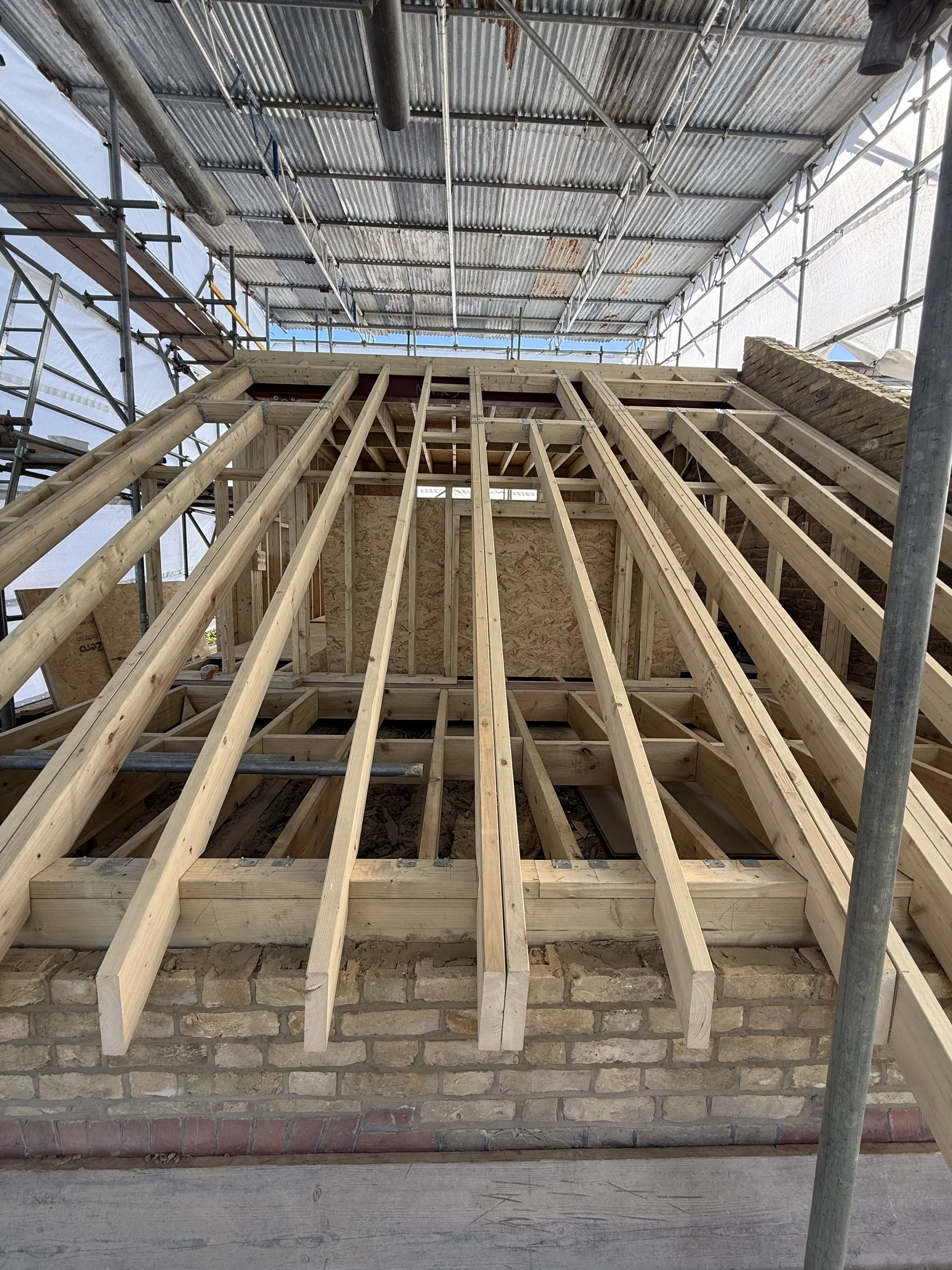 Construction site with a partially built wooden framing for a wall or roof, surrounded by metal scaffolding and a corrugated metal roof ceiling.