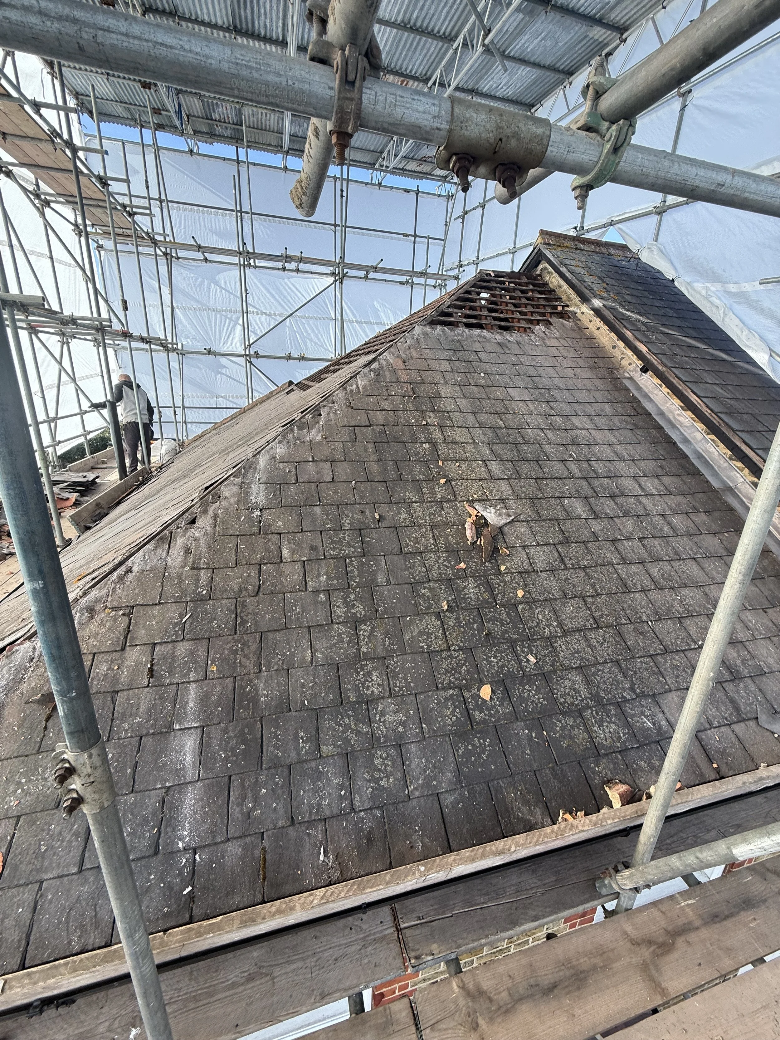Scaffolding around a house rooftop with roof tiles and a chimney, with a worker in the background.