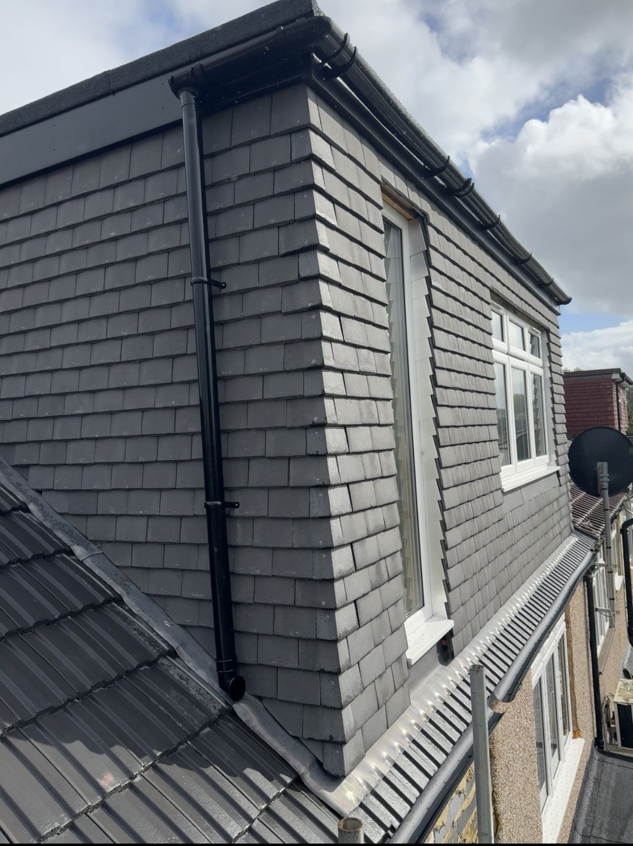 Photo of a house's roof showing gray shingle siding, a black rain gutter, and a black downspout under cloudy sky.