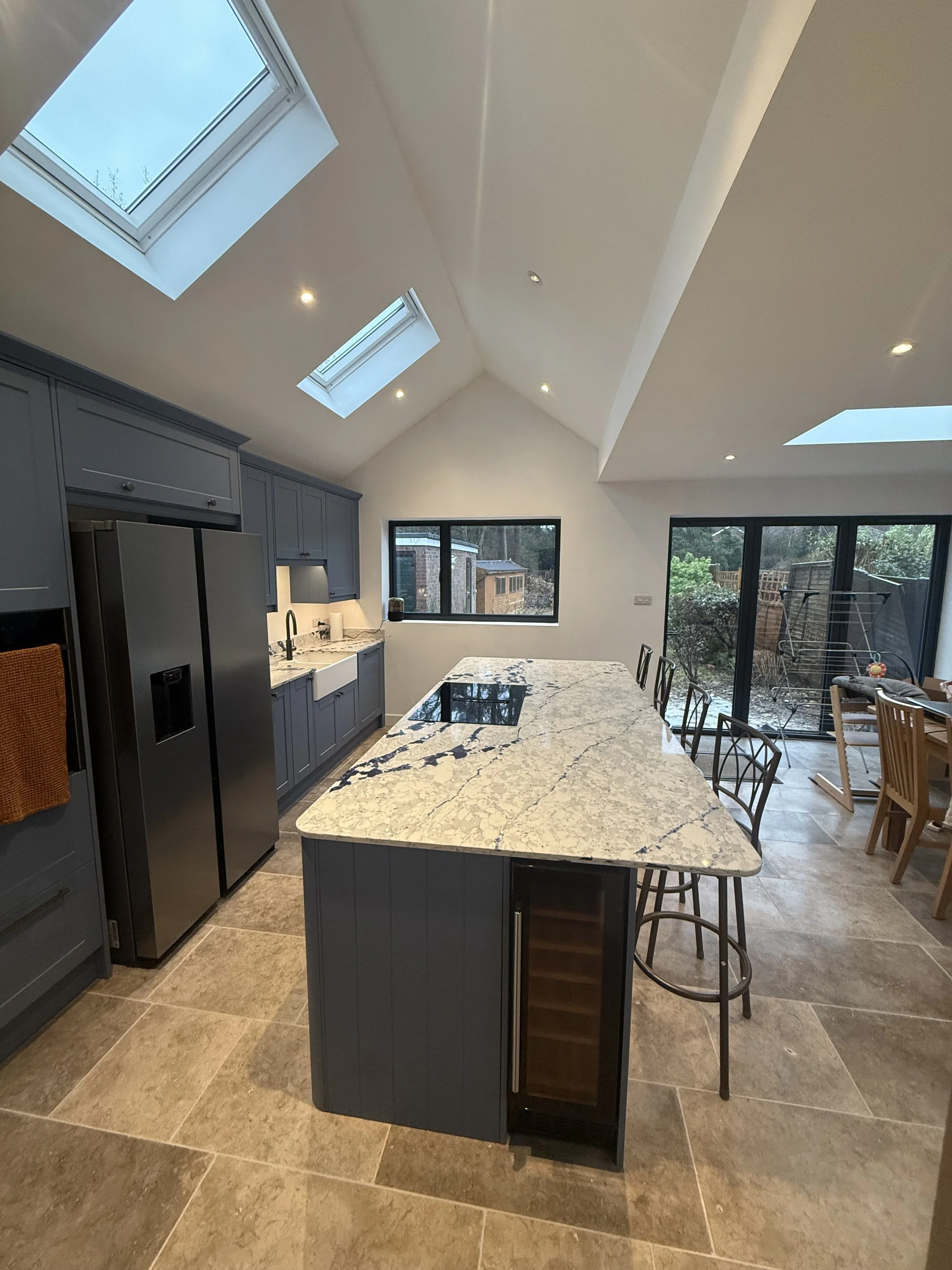 A modern kitchen with gray cabinetry, a large marble island, a stainless steel refrigerator, and skylights in a sloped ceiling. There are large windows and glass sliding doors showing a backyard with trees and a wooden fence.