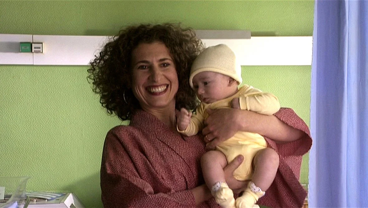 A woman with curly hair smiling while holding a baby dressed in yellow with a matching hat, in a room with green walls.