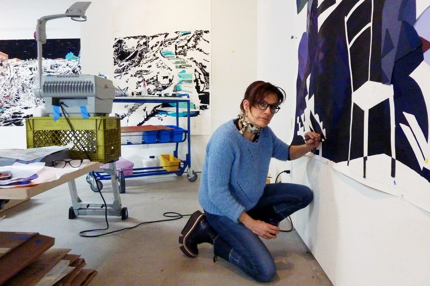 A woman wearing glasses, a blue sweater, and jeans is crouching and painting or drawing on a large abstract black and white artwork on a gallery wall. The room contains art supplies, a rolling cart with various items, and artwork on the walls.