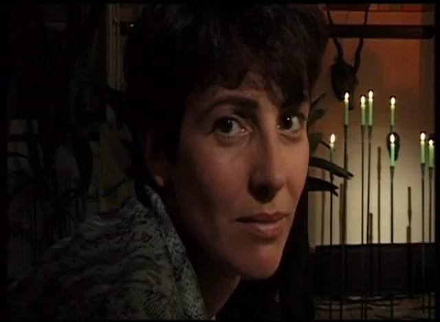 Person with short, dark hair looking over their shoulder in a dimly lit room with candles in the background.