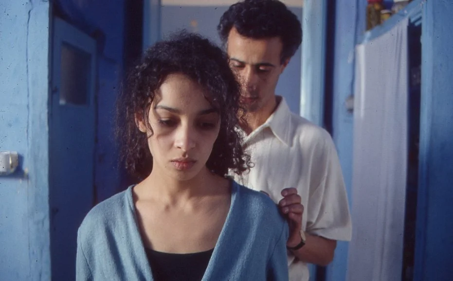 A woman with curly dark hair looking down with a somber expression, and a man with dark curly hair standing behind her, gently touching her shoulder in a blue hallway.
