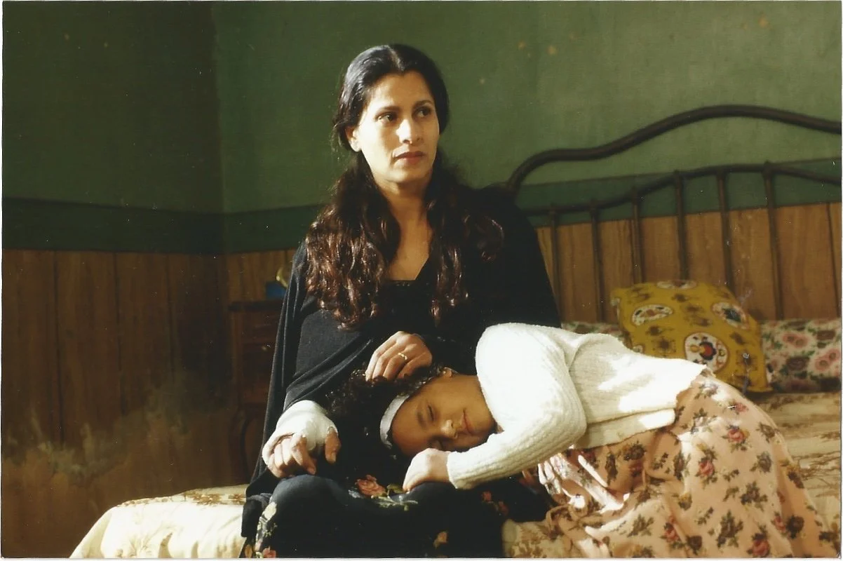 A woman with long brown hair sitting on a bed, with a young girl lying across her lap with her head resting on her arm.