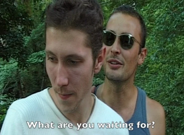 Two men outdoors, one wearing sunglasses and a tank top, the other wearing a white shirt, in a forested area.
